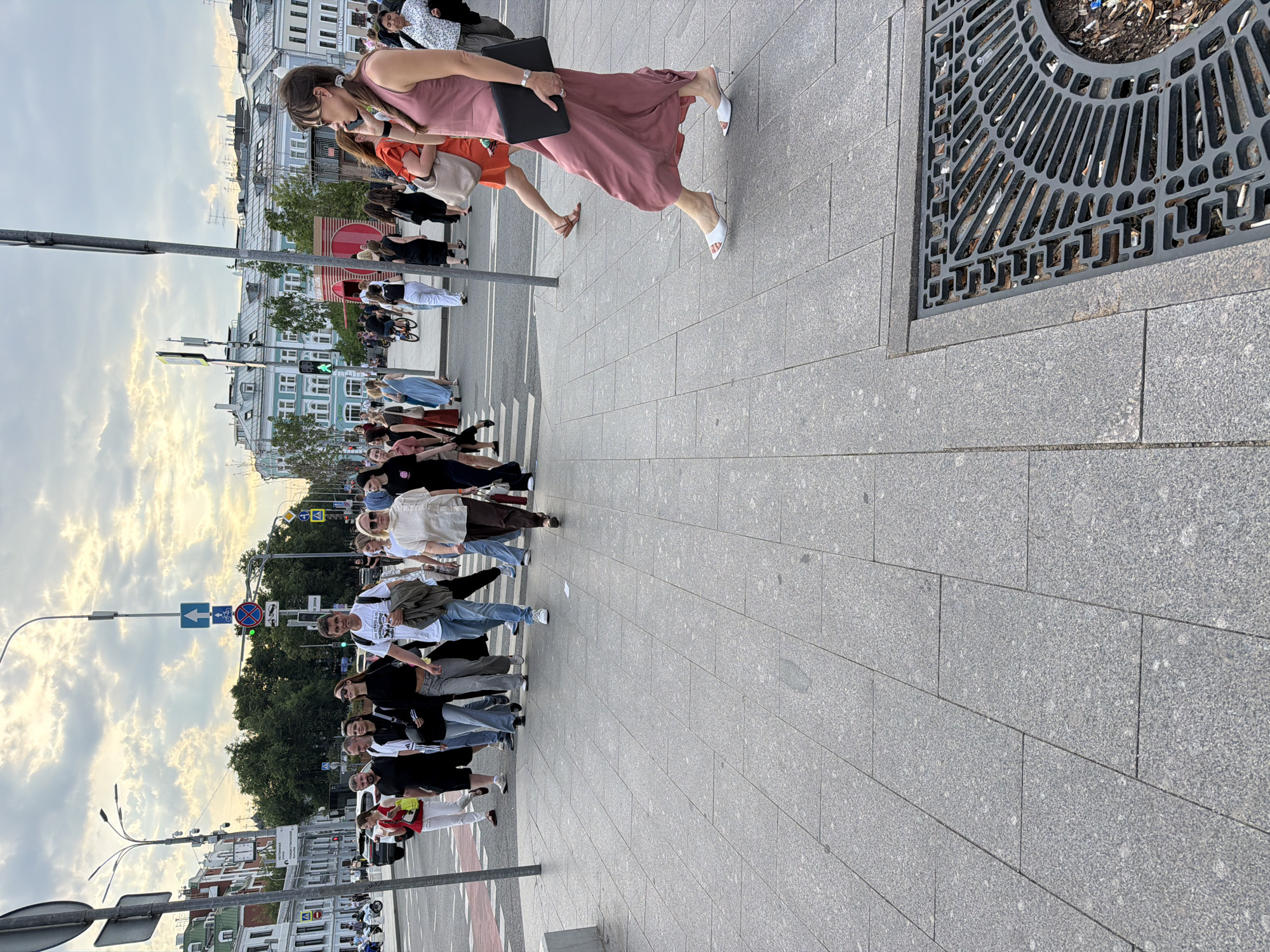The image depicts a street scene in Moscow, Russia, likely during daylight hours given the illumination. A group of pedestrians is crossing a street at a crosswalk. On the right side of the frame, a woman in a long, pink dress and white heeled sandals walks on a granite-tiled sidewalk, holding a black folder. Further back, other pedestrians are visible, including a man in a white t-shirt and jeans, a woman in a light-colored top and brown pants, and individuals dressed in casual attire. In the background, buildings with architectural details characteristic of Moscow can be seen, along with streetlights, traffic signals, and trees. The sky is cloudy, suggesting a diffused light. A storm drain grate with debris is also visible on the sidewalk in the foreground.