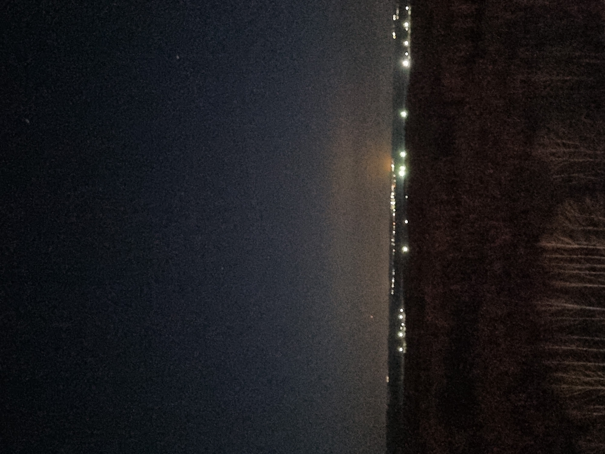 The image shows a dark night scene. The left side of the frame is dominated by a dark, grainy expanse likely representing the night sky. Along the right edge of the frame, a dark mass appears, possibly representing land. A line of faint lights is visible between the dark sky and the dark land mass. The grainy quality of the image suggests low lighting conditions or high ISO settings on the camera. The location and city name are not determinable from this image alone.
