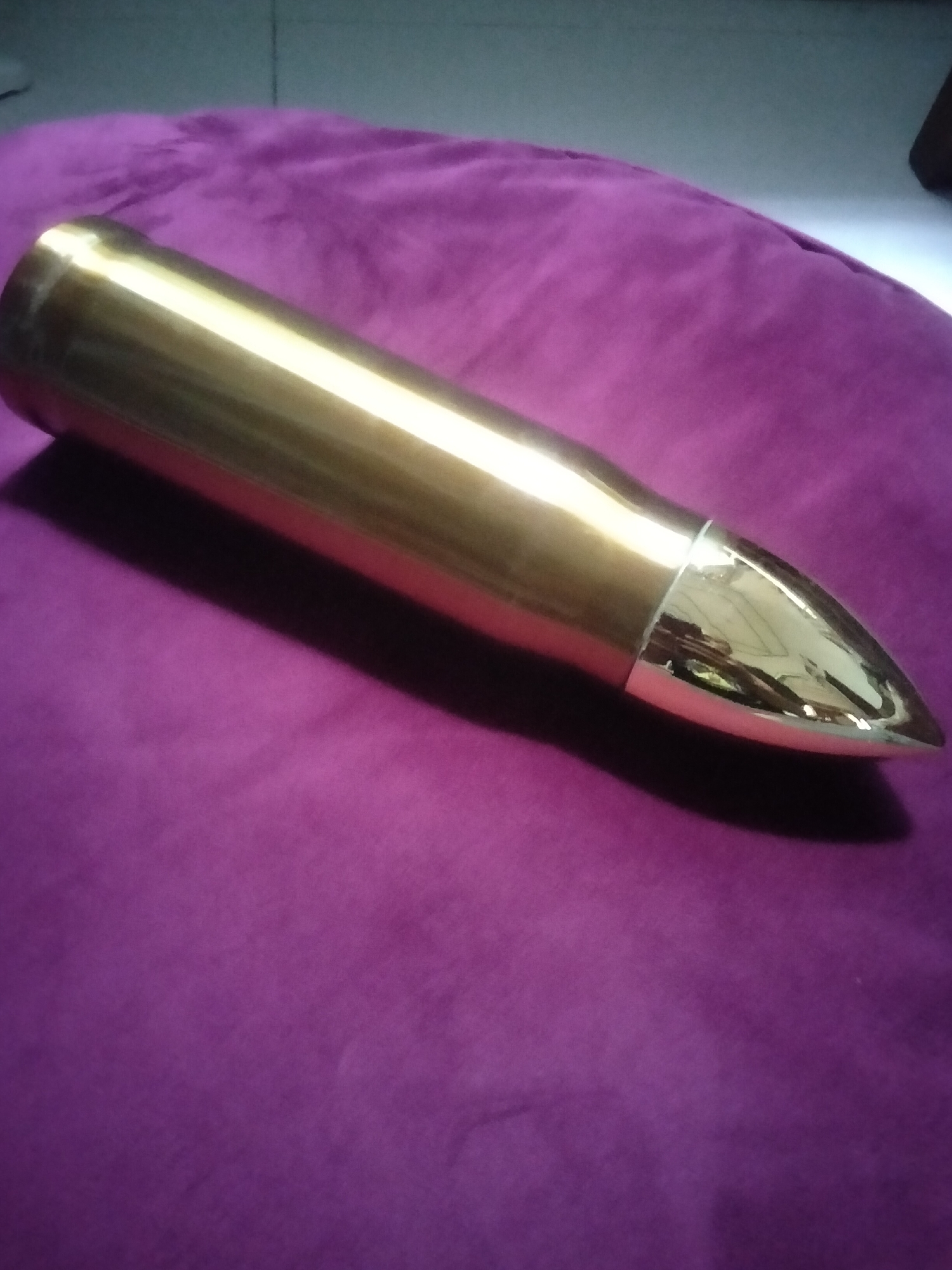 A polished, golden, bullet-shaped object lies on a purple, textured surface. The object is long and cylindrical, tapering to a pointed tip. The surface appears to be fabric, possibly velvet or a similar material, with visible creases and folds. The lighting suggests an indoor environment, with reflections visible on the shiny surface of the object. There is no indication of the object's location in Lhokseumawe, Indonesia, or any interaction between people and the object. The scene is static and focused on the object and its immediate surroundings.