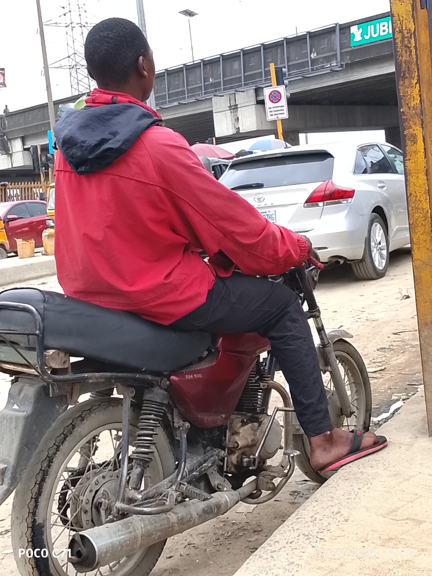 In Lekki, Nigeria, a person is seated on a red motorcycle, facing away from the camera. The individual is wearing a red jacket with a dark blue hood and black pants. The motorcycle is positioned on a dirt surface, with a curb visible in the foreground. In the background, there is a silver car, a yellow and red vehicle, and a traffic light with a "no stopping, no parking, no waiting" sign. An elevated road or bridge structure is also visible overhead, with a green sign displaying "JUBILEE" and a stylized image. The sky is overcast. The timestamp "07/26/2026 13:02" is overlaid on the lower right of the image, and "POCO C71" is on the lower left.