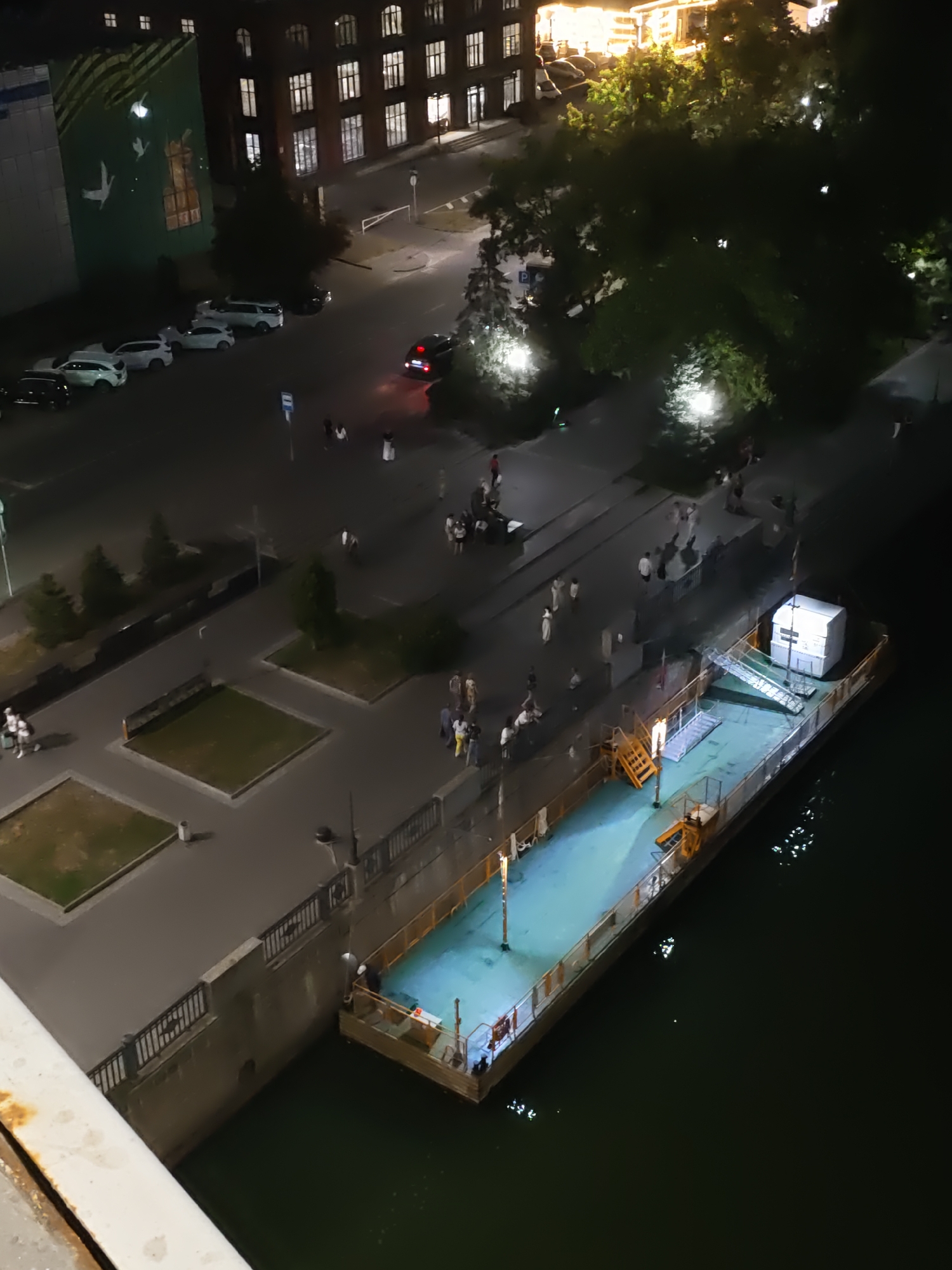 The image depicts a nighttime scene in Rostov-on-Don, Russia, likely near a waterfront area. On the left, a multi-story building with lit windows is visible, adjacent to a large mural of white birds against a green background. Below this building, a street is lined with parked cars, and a few pedestrians are present.

In the center and right of the frame, a wide paved walkway runs along a concrete embankment. This walkway is populated by numerous individuals, some walking in small groups, others standing. Lampposts illuminate parts of the walkway and surrounding trees.

Dominating the lower right portion of the image is a long, flat-bottomed barge or a similar vessel docked alongside the embankment. The deck of the barge is lit, revealing a light blue surface and some structures on it, including a white box-like building at the far end and staircases leading to the deck. The water, dark and reflective, surrounds the barge, with ripples and the lights of the barge creating shimmering patterns. A section of a bridge railing is visible in the foreground, suggesting the photograph was taken from a higher vantage point. The overall atmosphere is one of urban activity at night, with a mix of residential or commercial buildings, public spaces, and maritime elements.