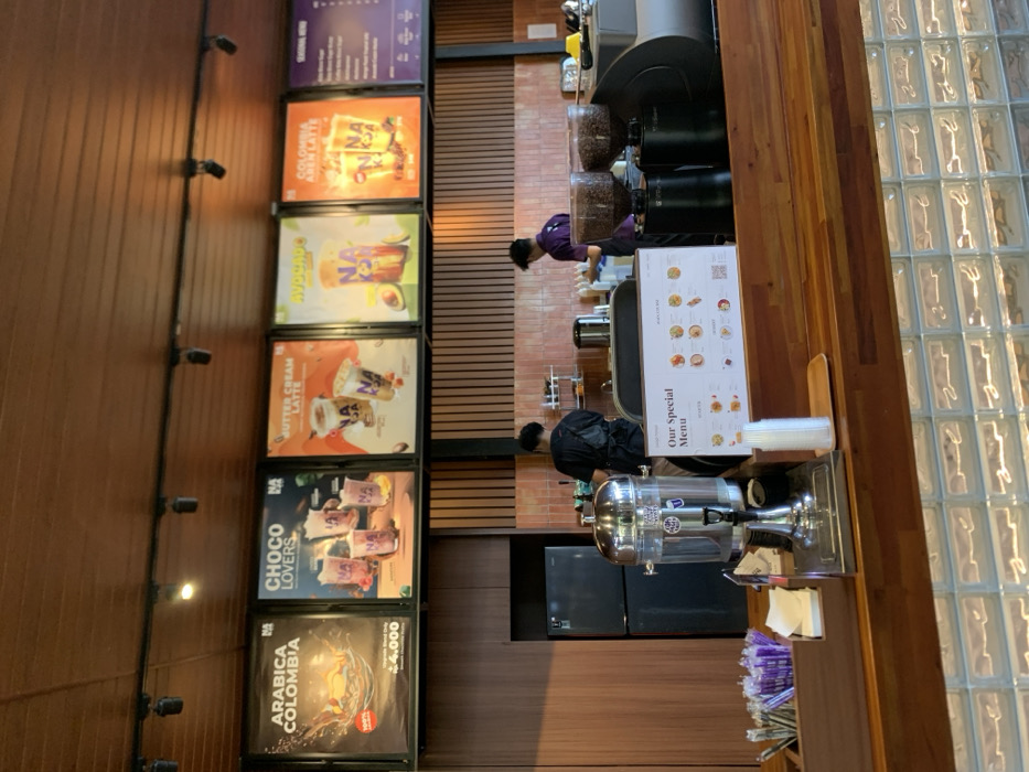 The scene is a cafe located at the coordinates -7.957, 112.622, in the city of Malang, Indonesia. Two men are standing behind the counter. One is facing the camera, wearing a black shirt, the other is facing away from the camera, wearing a purple shirt. There are several glass containers behind the counter with unknown liquids in them, along with a black, stainless steel coffee grinder. On the counter, there is a large, stainless steel container with clear liquid and a "Our Special Menu" sign. There are two smaller stainless steel coffee grinders and a large, stainless steel espresso machine. There is a large, black, metal refrigerator behind the counter. Five framed posters are on the wall behind the counter, each advertising different types of coffee or tea. The counter is made of wood. Below the counter there is a glass brick wall. There are some drinking straws in a container next to the counter.