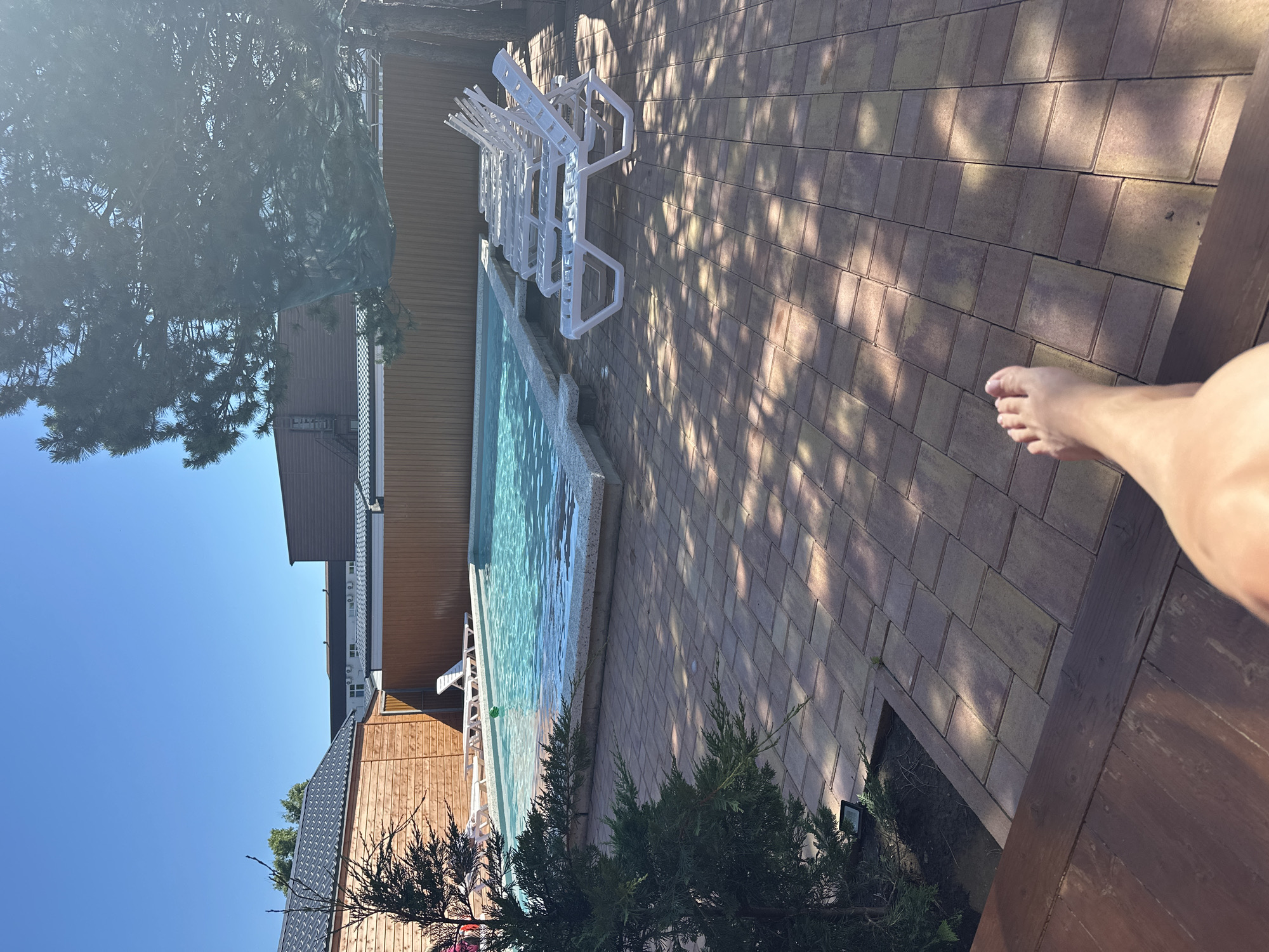 The image depicts a sunny day at an outdoor swimming pool area. The pool is filled with clear blue water and surrounded by a paved patio. Several white lounge chairs are arranged neatly on the patio, facing the pool. A wooden fence encloses the area, and some trees and buildings are visible in the background. A person's bare foot is prominently featured in the foreground, suggesting they are relaxing by the pool. The scene appears to be a resort or a private residence in Anapa, Russia, given the context.