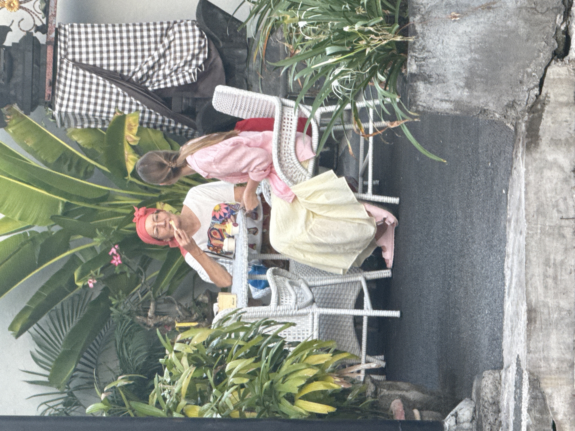 In Denpasar, Indonesia, an eye-level shot shows a woman with a red headscarf seated at a white wicker table outdoors, eating with chopsticks. A young girl with long hair stands next to her, looking at the table. Lush green plants and flowers surround them, with a checkered cloth and assorted objects visible behind them. The setting appears to be a street-side cafe or restaurant. The ground is paved, and a weathered wall runs along the edge of the scene.