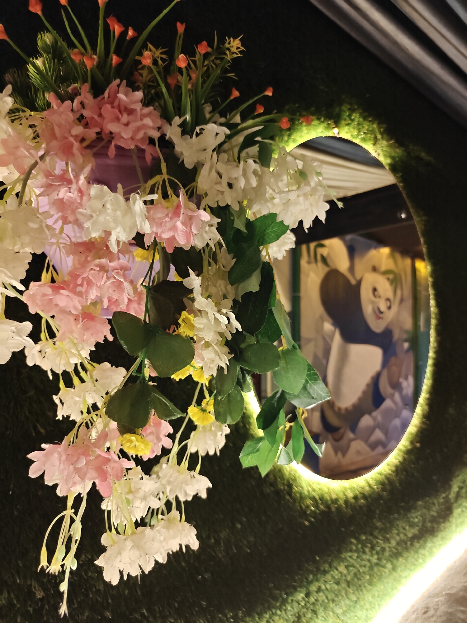 This image shows a close-up of artificial flowers in various shades of pink, white, and yellow, with green leaves, suspended in front of a circular mirror. The mirror reflects a mural of a cartoon panda character. The mirror is framed by a textured green surface, possibly artificial grass, which is illuminated by a soft yellow light. The scene appears to be indoors, potentially a restaurant or a themed establishment. There are no people visible in the image. The overall impression is one of decorative artificial foliage and a whimsical reflected image. The location in Pokhara, Nepal, is not identifiable from the visual information provided in the image itself.