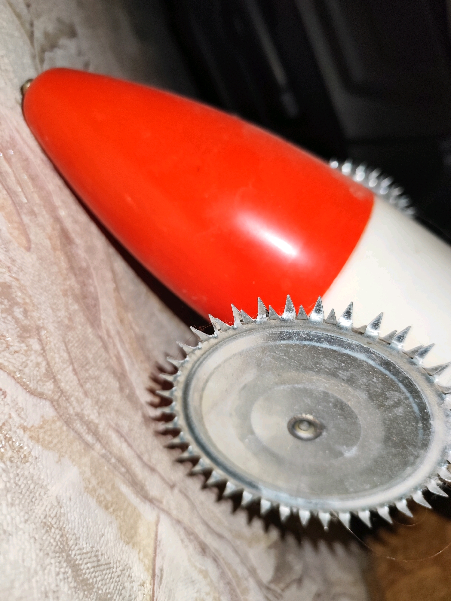 The image is a close-up shot of a vintage electric hand mixer. The mixer has a red and white plastic body and a metal whisk attachment with sharp teeth. The mixer is resting on a textured surface, possibly a tablecloth or placemat, with a light beige and brown marbled pattern. The background is dark and out of focus. There is no discernible location in the city of Balqaş, Kazakhstan.