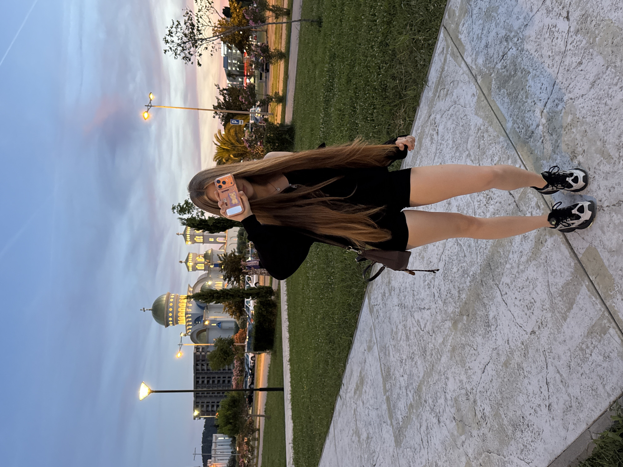 A young woman with long, brown hair stands on a paved walkway in front of a lawn. She is wearing a black top, black shorts, and black and white athletic shoes. She is holding a smartphone in her right hand, obscuring her face. In the background, a large, white, domed building with gold accents, possibly a church, is visible. The sky is a dusky blue with hints of pink and orange, suggesting it is either dawn or dusk. Streetlights are visible, and trees and parked cars are also present in the background, indicating an urban setting. The scene appears to be in the city of Bar, Montenegro, given the architectural style of the building and the overall environment.