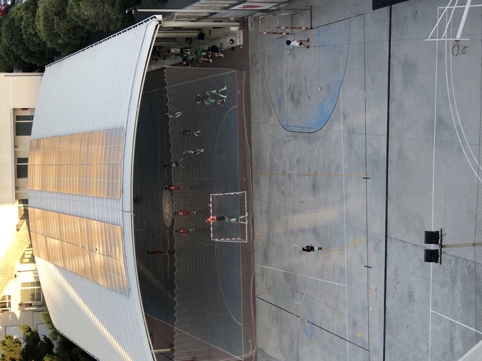 The image shows an outdoor sports court with a covered section featuring solar panels on the roof, located in the city of València, Spain. The court appears to be a futsal or basketball court, with painted lines and markings on the concrete surface. There are two small goals visible on the court. In the covered area, a group of young people, dressed in athletic wear, are playing futsal. One individual in a green and white uniform is in goal. Several other individuals, some in red and white uniforms, are scattered on the court. Outside the covered area, a single individual in a white shirt and black shorts is walking across the court. There are also spectators sitting on a bench in the covered area. In the background, a building with multiple stories is visible, along with trees and a street. The lighting suggests it is either early morning or late afternoon, with sunlight casting long shadows.