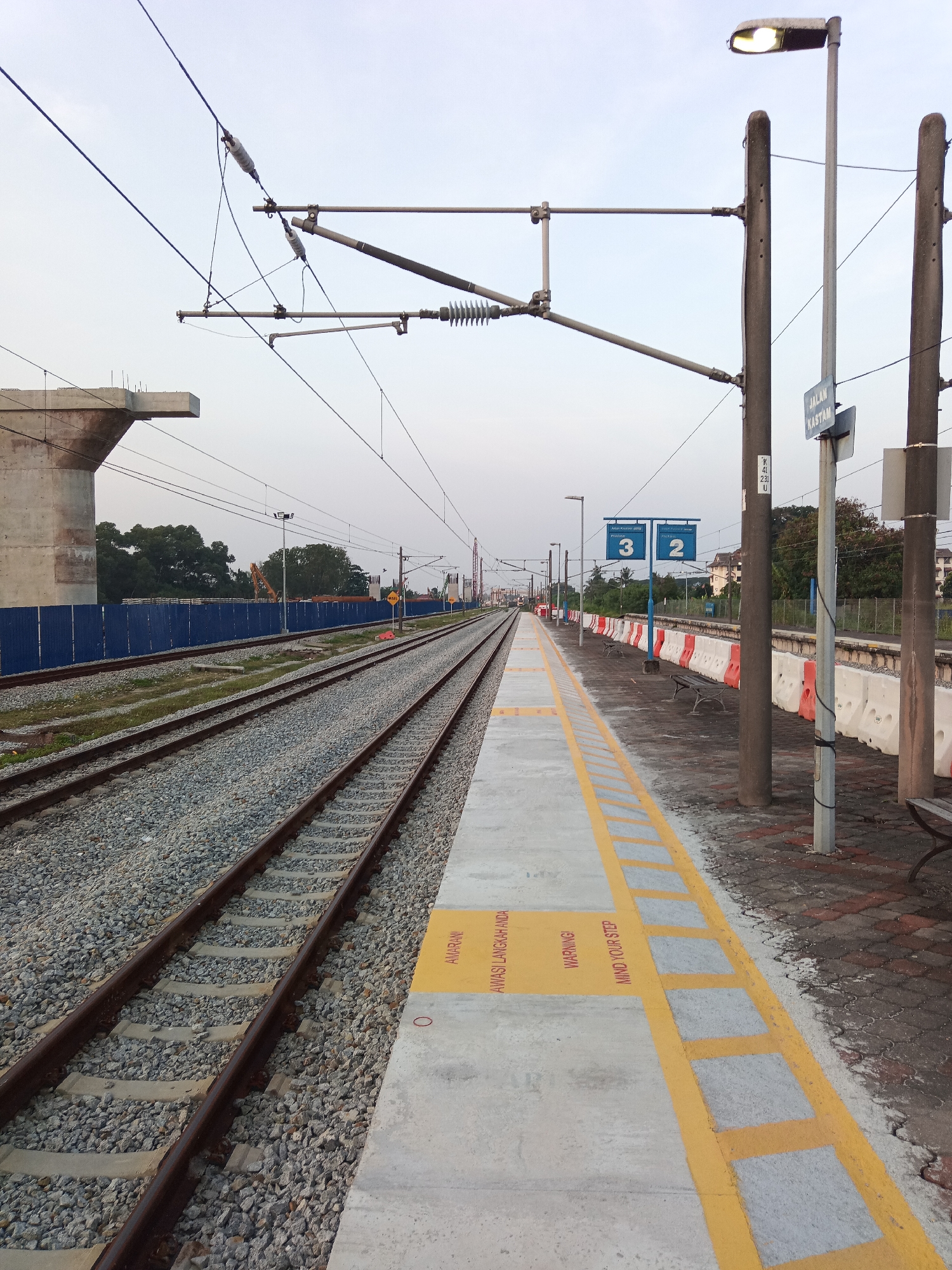 This is a medium-distance, eye-level, outdoor, daytime photograph of a train station in Klang City, Malaysia. The scene is dominated by multiple sets of train tracks, a concrete platform with yellow safety markings, and overhead electrical lines. In the foreground, a paved platform extends to the right, featuring yellow lines and warning text in English and Malay. Benches are visible on the platform, along with concrete and orange barriers separating the platform from the tracks.
In the middle ground, two sets of gravel-filled train tracks run parallel into the distance. A blue construction barrier lines the left side of the tracks, behind which trees and construction equipment, including a yellow excavator, are partially visible. Overhead, a complex network of electrical lines and support poles are strung across the sky.
In the background, more train tracks and overhead lines continue into the horizon. A large, unfinished concrete structure is visible on the far left, suggesting ongoing construction. A street lamp illuminates the platform on the right. A sign identifying "JALAN KASTAM" (Kastam Road) is affixed to a pole. The sky is overcast and pale, indicating early morning or late afternoon light. There are no people visible in the image.