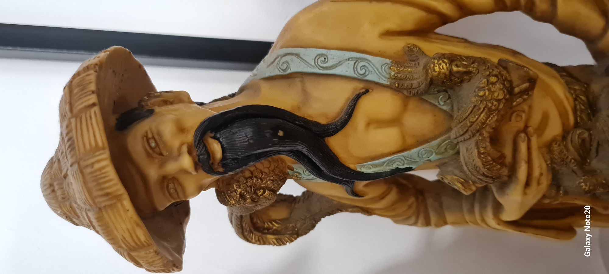 This is a close-up, eye-level, medium shot of a statue of an East Asian man wearing a straw hat, a tan robe, and a blue sash adorned with swirling patterns. The statue has a long, black beard that splits into three strands. The man's arms are positioned as if carrying something, and a golden, feathered object is draped over his left shoulder. The statue is made of a light brown material with golden accents. The background is a plain white wall with a dark vertical line on the right side. The bottom left corner of the image has a watermark that reads "Galaxy Note20". The statue is positioned indoors, likely in a home or shop, and its specific location within the city of Holon, Israel, cannot be determined from the image alone.