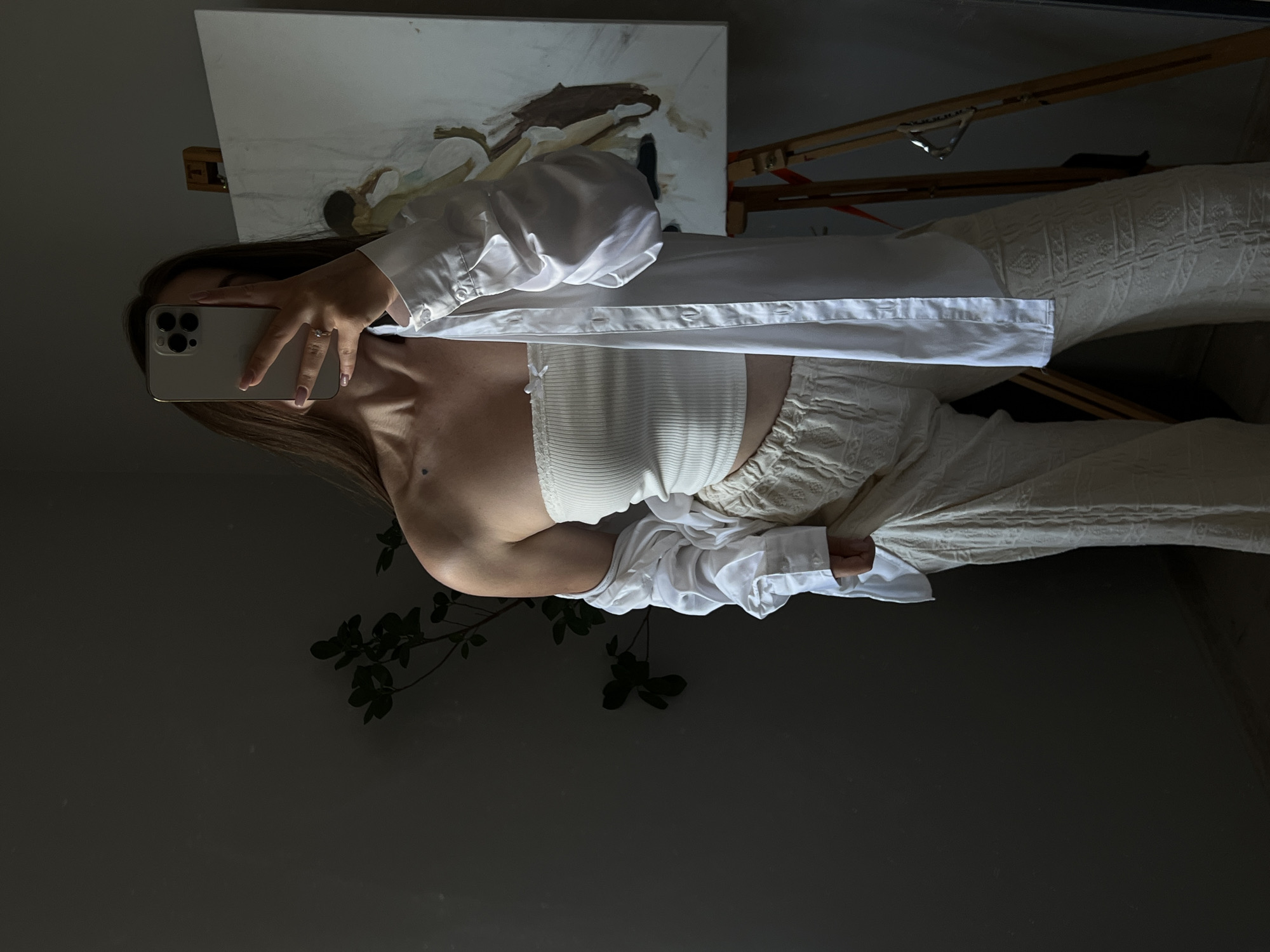 The image captures a person taking a mirror selfie in what appears to be a room with an easel and a painting. The person is wearing a white ribbed tube top and a white unbuttoned oversized shirt, with cream-colored pants. Their right hand is holding a smartphone, and a ring is visible on their left hand. Behind them, a painting is propped on an easel. The scene is dimly lit, suggesting it might be evening or the room has limited natural light. A plant with dark green leaves is visible in the foreground, casting shadows on the dark wall. The overall mood seems relaxed and personal, typical of a casual selfie. The image is oriented vertically, with the top of the person's head towards the left side of the frame. There is no visible text in the image.