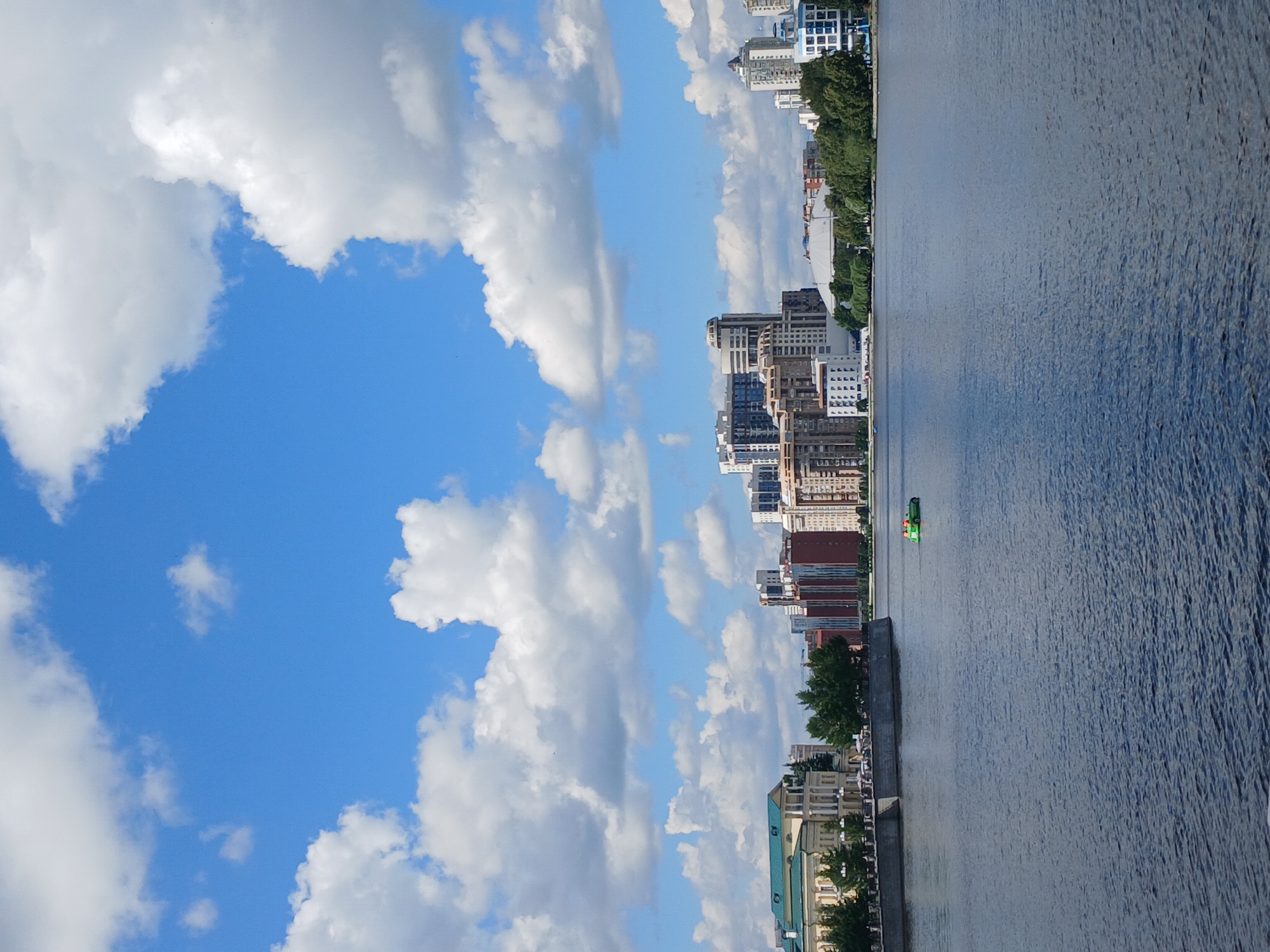 This media file depicts a scenic view of a city skyline alongside a body of water, likely a river, on a bright, cloudy day. The scene is oriented vertically, suggesting the original photo was taken in portrait mode or has been rotated.
On the left side of the image, a vibrant blue sky is filled with large, puffy white clouds. The right side of the frame showcases a cityscape with a variety of buildings, ranging from modern high-rises to older structures, lining the waterfront. Lush green trees are interspersed among the buildings, providing a natural contrast to the urban architecture.
The water is calm, with gentle ripples on its surface, reflecting the sky and surrounding structures. A small, bright green boat is visible on the water, adding a touch of color and suggesting movement or activity. Along the edge of the water, there's a concrete embankment or promenade, with some pedestrians possibly present, though they are not clearly discernible.
The overall atmosphere appears peaceful and pleasant, indicative of a daytime scene. The lighting suggests it is likely late morning or afternoon. The presence of diverse architectural styles hints at a city with a history and ongoing development. While specific location cues within the image are limited to the general urban and waterfront setting, the provided context of Yekaterinburg, Russia, helps place the scene geographically. No discernible text is present in the image.