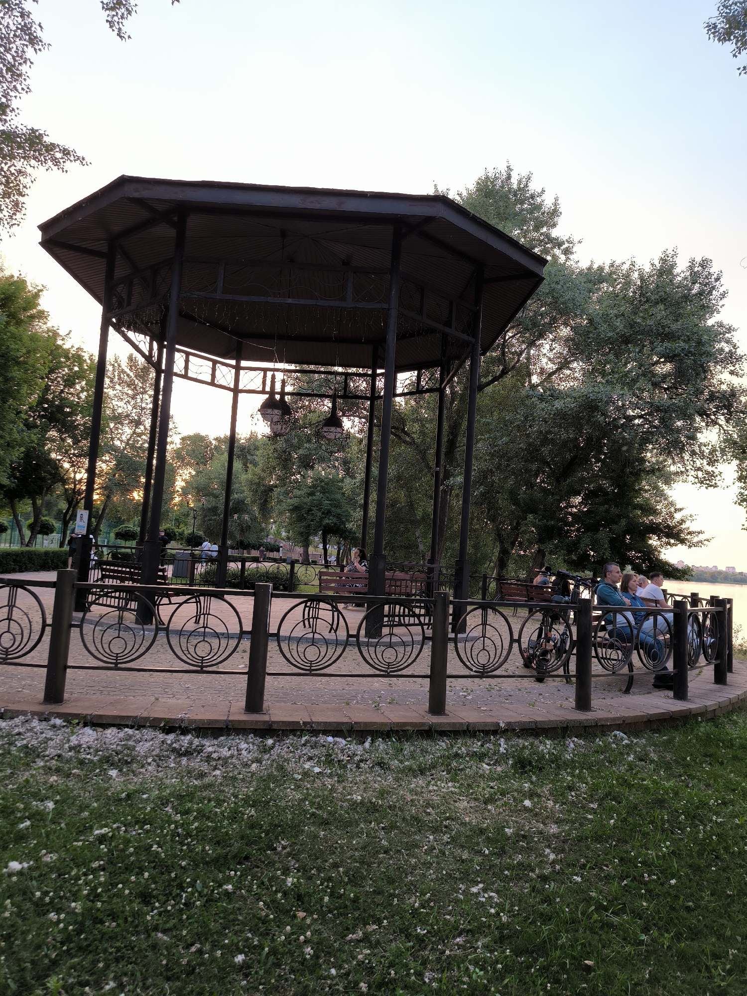 The image depicts a park scene with a large, dark-colored hexagonal gazebo at its center. The gazebo has a metal roof supported by dark pillars. Inside and around the gazebo, several people are seated on benches. In the foreground, a decorative metal fence with circular motifs separates the paved area from a grassy lawn littered with white fluffy material, possibly from cottonwood trees. Beyond the gazebo, trees and foliage are visible, with a hint of a body of water in the background, suggesting a park situated near a river or lake. The lighting suggests it is either early morning or late evening, with the sun casting a warm glow through the trees. A few bicycles are parked near the fence. The overall impression is a tranquil park setting.