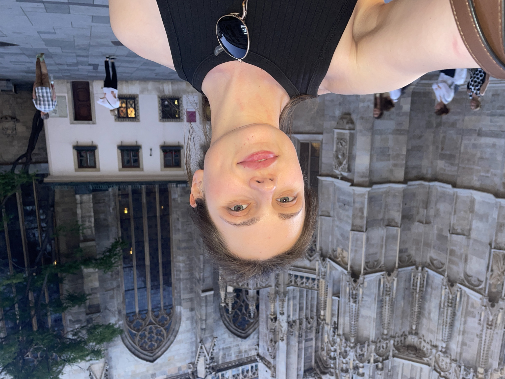 A person takes a selfie in front of the St. Stephen's Cathedral (Stephansdom) in Vienna, Austria. The individual, wearing a black tank top with sunglasses, is positioned with the cathedral's detailed facade prominently in the background. Other buildings and people are visible in the distance.