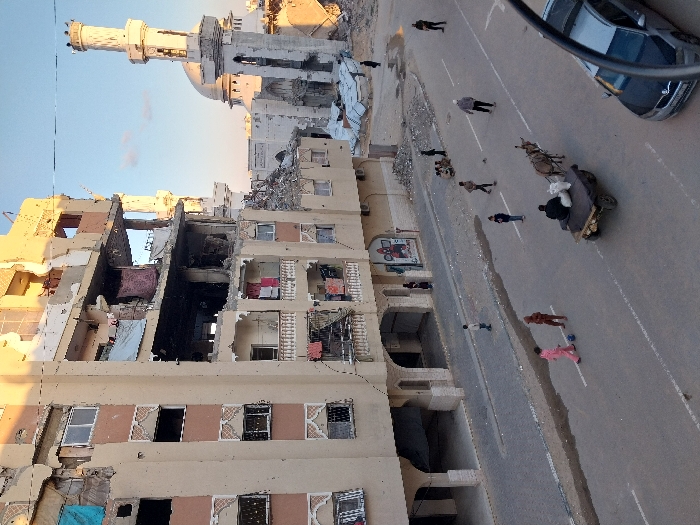 The image depicts a street scene in Khan Yunis, Palestinian Territory, during late afternoon or early morning. A multi-story residential building on the left shows significant structural damage, with upper floors partially collapsed, exposed interiors, missing windows, and damaged balconies. Debris is visible within and around the structure. To the right, a mosque with a distinct minaret and dome is also damaged, with rubble present at its base and a partially destroyed minaret top. The paved street below features white lane markings and patches of debris or moisture. Numerous individuals populate the street: two children in pink and brown clothing play with a ball in the road, a man sits on a donkey-drawn cart moving away from the viewer, and several other adults and children walk or stand along the street. A silver sedan is partially visible on the far right. The scene is illuminated by warm, low-angle sunlight, casting long shadows.