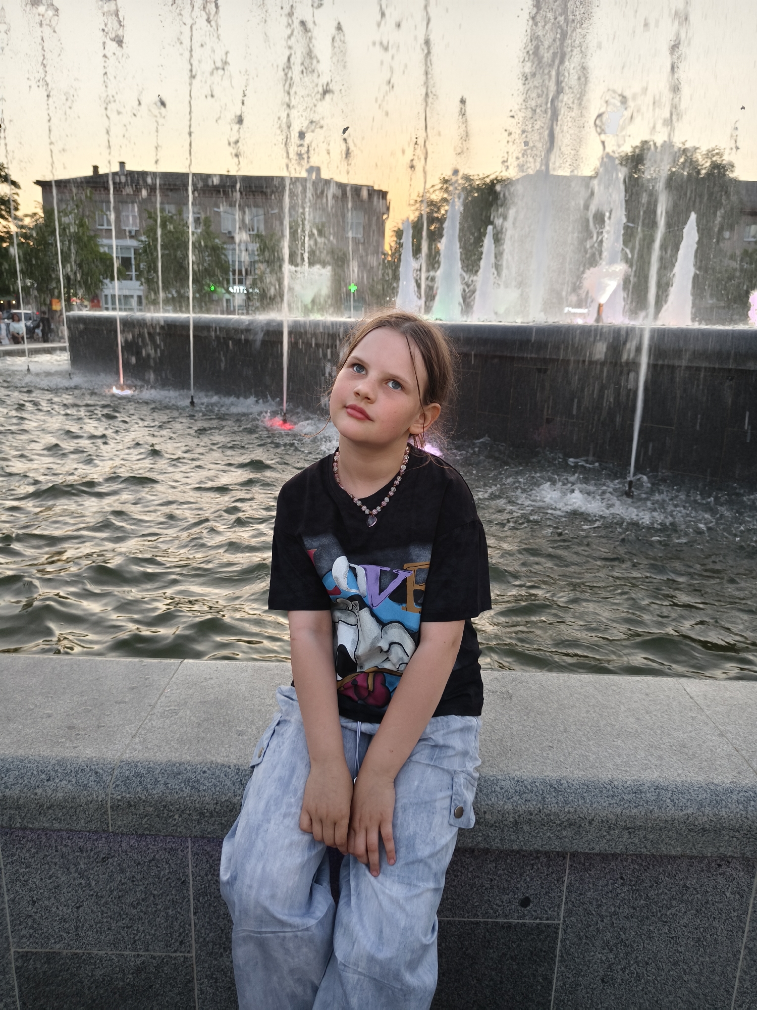 A young girl sits on a gray concrete ledge in front of a large fountain. The girl is wearing a black t-shirt with a cartoon graphic, light blue pants, and a beaded necklace. She is looking up and to the left. The fountain has multiple water jets spraying upwards, with a building visible in the background. The time of day appears to be late afternoon or early evening, judging by the lighting. The location is a city, but the specific city is unknown.