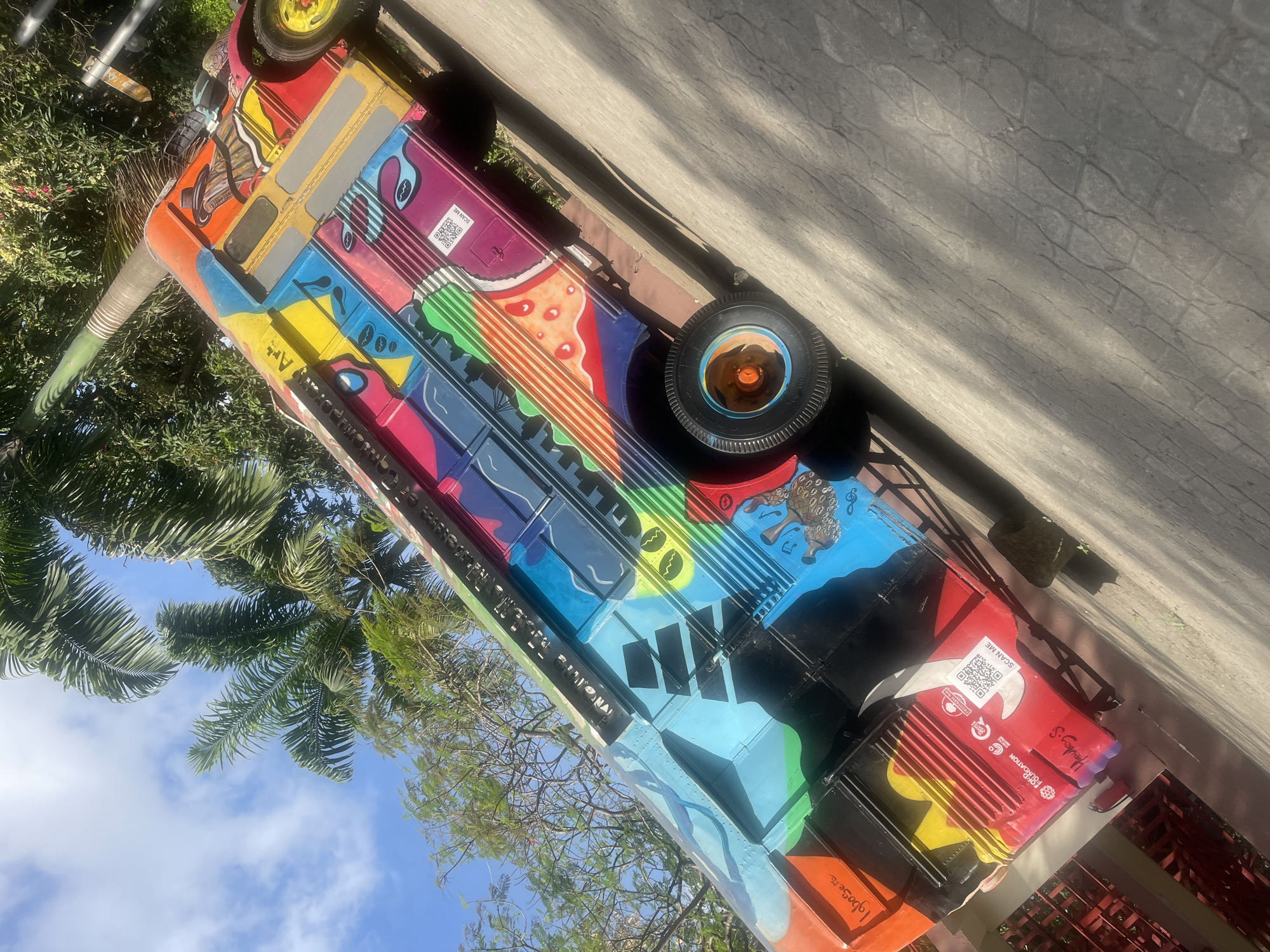 The image shows a vibrantly painted bus against a backdrop of palm trees and a partly cloudy sky, possibly located outdoors in a tropical or subtropical climate. The bus is adorned with colorful artwork featuring geometric shapes, musical notes, and QR codes. The bus appears to be stationary, potentially on display as an art installation or repurposed vehicle. It has multiple sponsors like IgboSere, Freedom Foundation, and Hawley S. The words "Museum of Contemporary Art" appear along the side of the bus, further suggesting its artistic purpose. The overall impression is that of a public art piece blending cultural expression with repurposed utility.