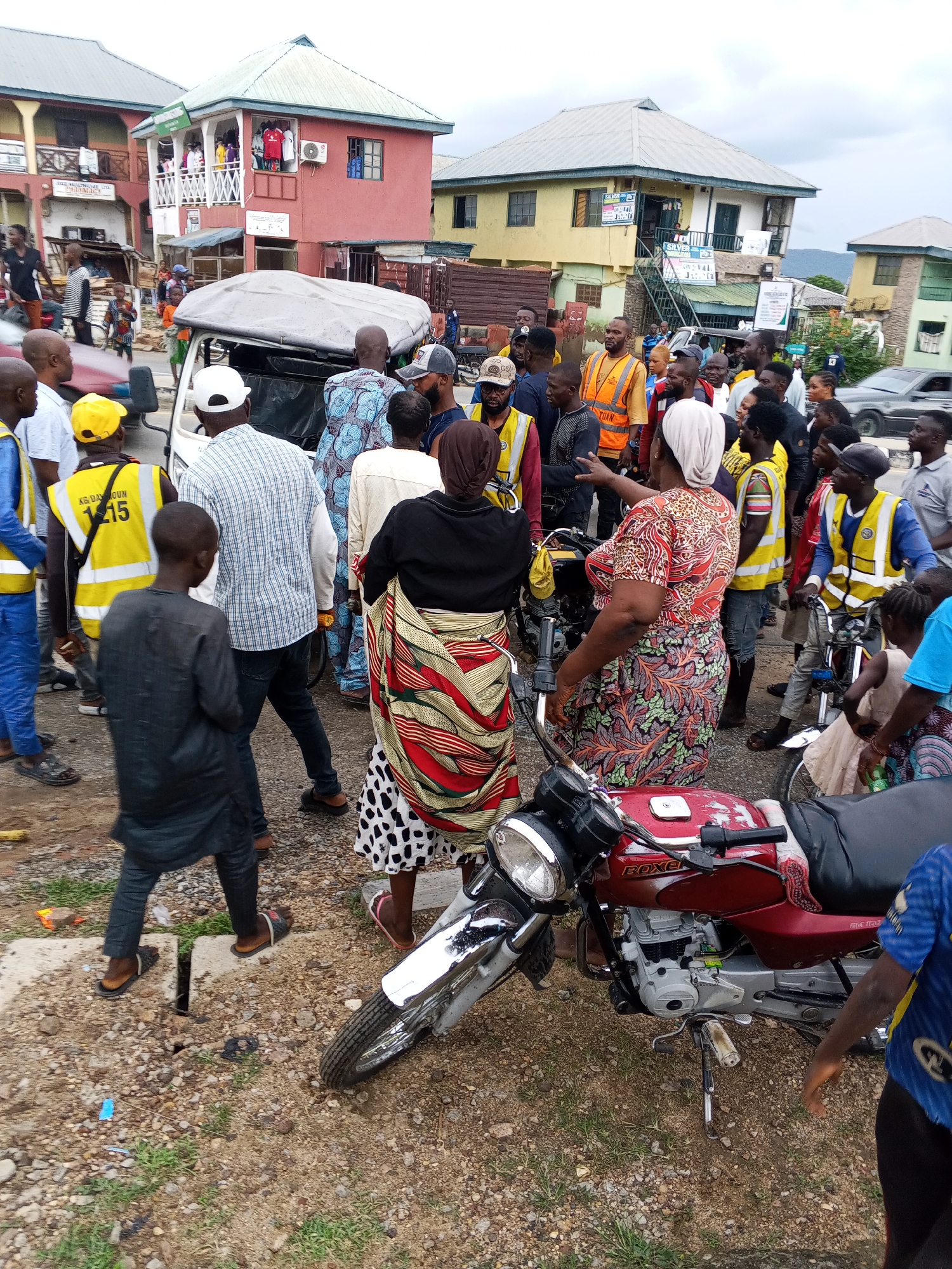 The image depicts a bustling street scene in Zango-ndaji, Nigeria, where a crowd has gathered around a damaged motorcycle and a white van. The atmosphere suggests a recent incident, possibly an accident, as indicated by the presence of several individuals wearing high-visibility vests, common for traffic control or emergency personnel.
In the foreground, a red motorcycle lies on its side, its front tire detached. A man in a blue t-shirt and black pants is standing next to it, appearing to inspect the damage. Behind him, a woman in a patterned dress and headscarf is gesturing towards the scene, possibly explaining what happened.
A white van, partially obscured by the crowd, is also visible. It appears to have its rear doors open. Several individuals are clustered around the van and the motorcycle, some looking concerned, others engaged in conversation. Many people in the crowd are wearing casual clothing, but a few are in bright orange or yellow vests, suggesting they are involved in managing the situation.
The background reveals buildings typical of a Nigerian urban environment, with colorful facades and multi-story structures. The street is paved with a mix of asphalt and gravel, and debris is scattered on the ground. The sky is overcast, indicating a cloudy day. The overall impression is one of a minor traffic incident that has drawn the attention of onlookers and potentially emergency responders.