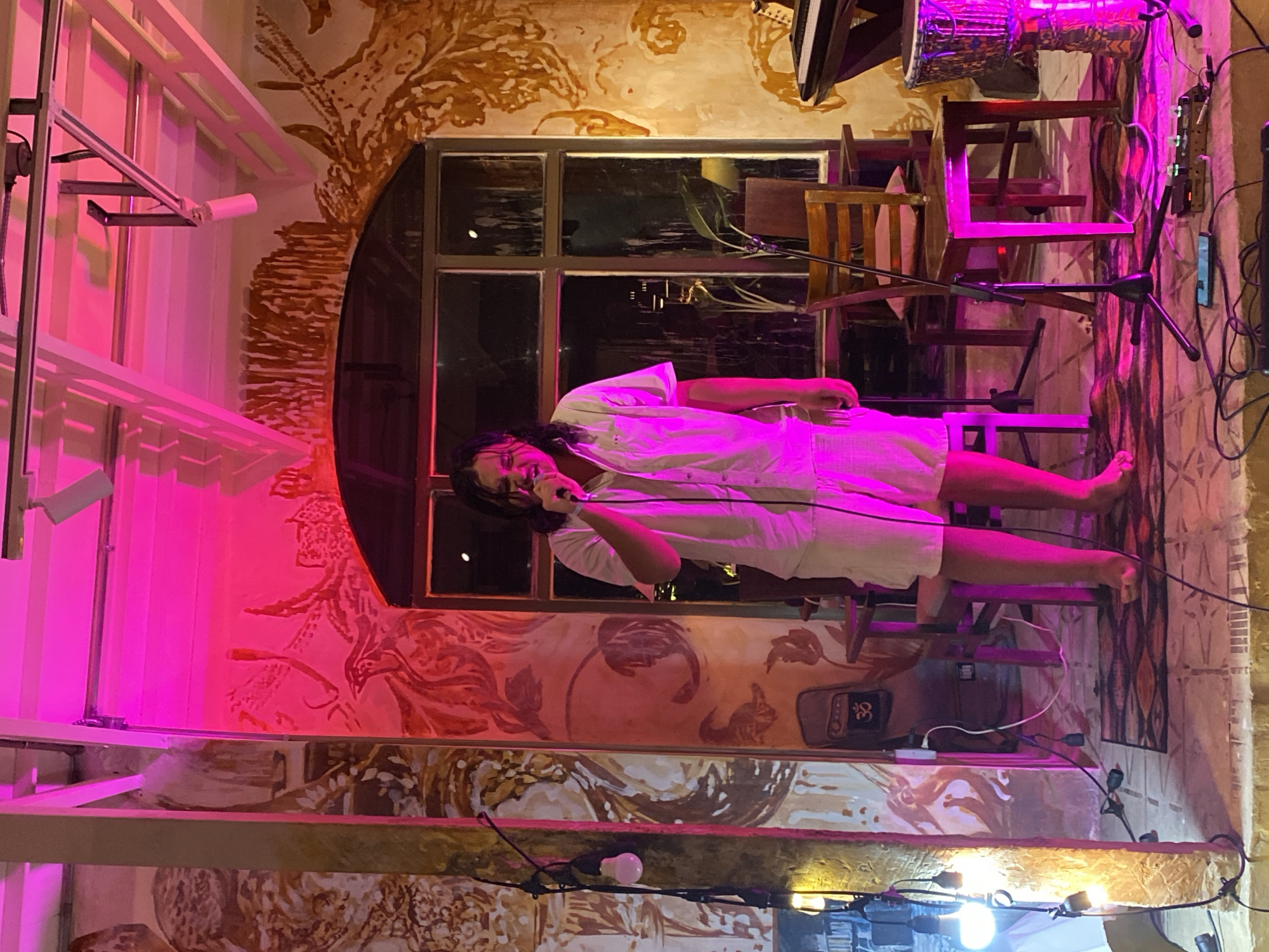 In this vertically oriented image, a person is singing into a microphone. They are dressed in a white, loose-fitting garment and appear to be on stage or performing in a venue. The background suggests an indoor setting with artistic murals painted on the walls, featuring abstract designs in earthy tones of brown and tan. There are also windows in the background that look out onto a dark, possibly nighttime scene.
The scene is illuminated by vibrant pink lighting on the left side, which casts a striking glow on the surroundings and the performer. Other lights, including strings of bare bulbs, are visible in the lower portion of the image. Several wooden chairs are stacked and arranged in the background, some of them also catching the pink light. A djembe drum is partially visible at the top, suggesting a musical performance. Electrical equipment, including a power strip and cables, is also present.
The performer's posture and the microphone indicate they are actively singing. The overall mood is somewhat intimate and atmospheric, enhanced by the colored lighting and the artistic backdrop. The time of day appears to be evening or night, given the darkness outside the windows. There is no visible text in the image.