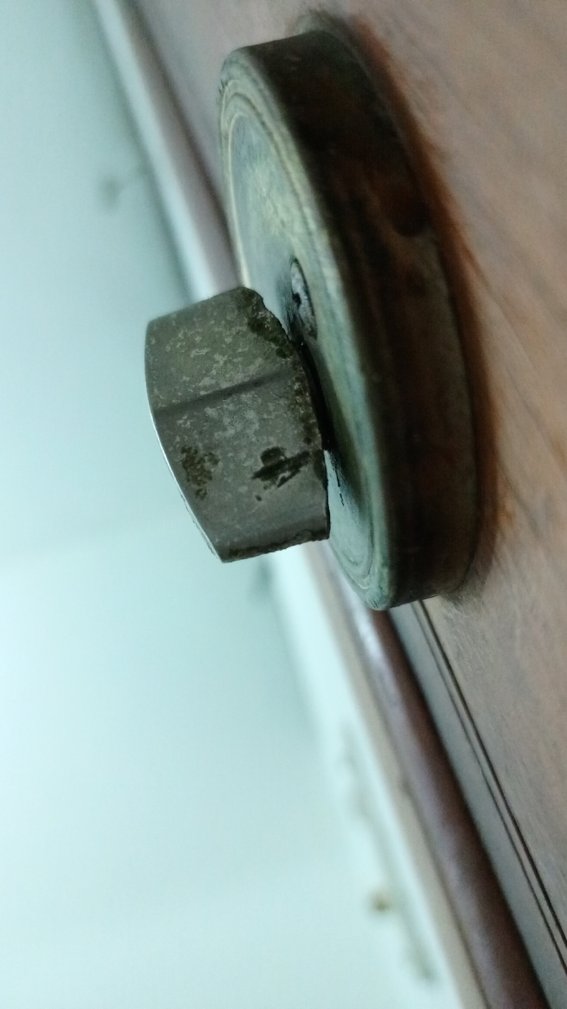 A close-up, slightly tilted image shows an antique-style metal knob attached to a wooden surface, likely a door or cabinet. The knob features a round, disc-shaped base and a square, protruding handle. The metal exhibits signs of age and wear, with a tarnished, textured surface. The wooden surface has a dark, polished finish with visible grain. The background is a blurry, light-colored wall. Location information is not determinable based on this image.