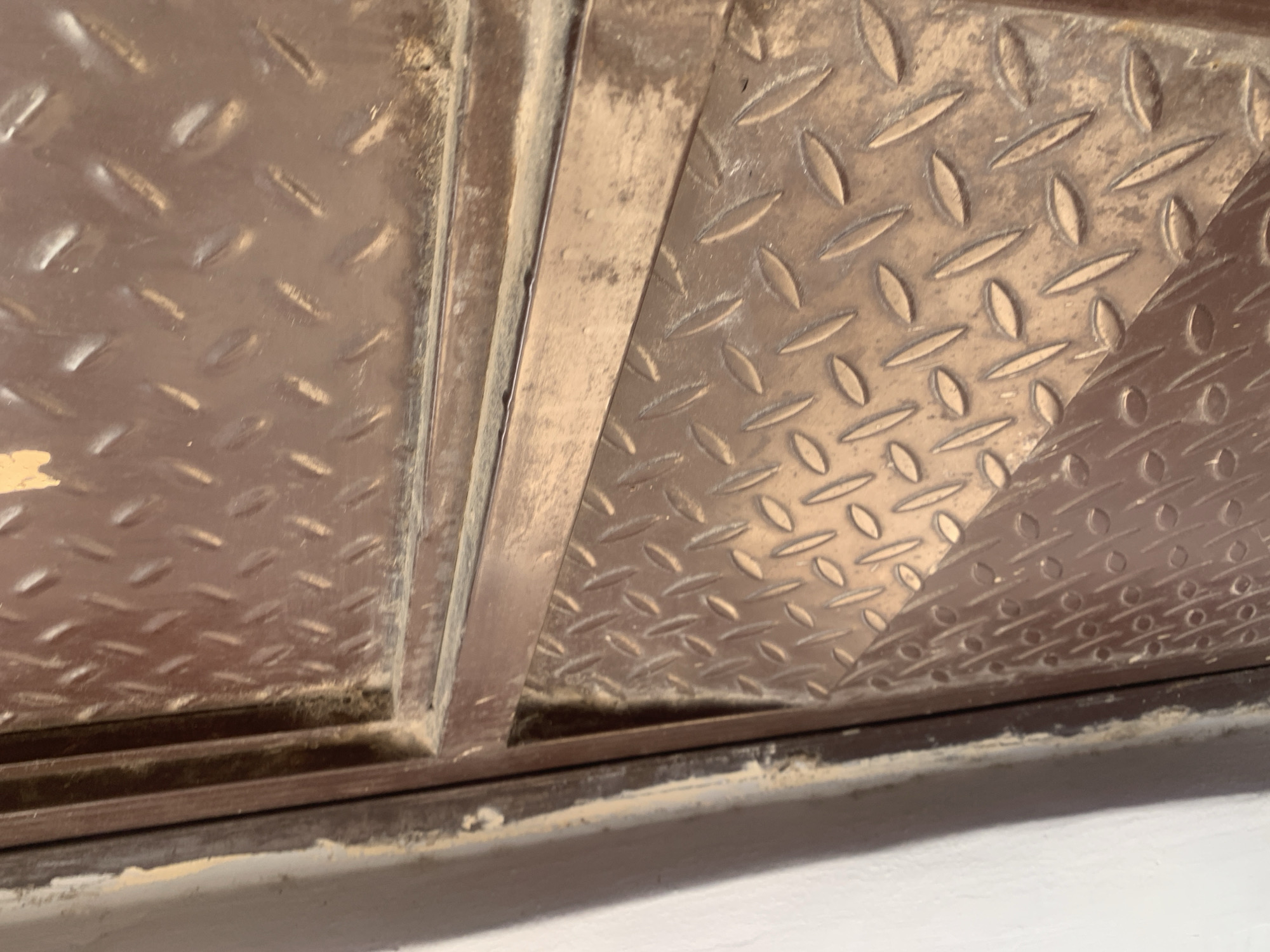 The image shows a close-up view of a metal door or gate with a diamond plate pattern. The metal is a reddish-brown color and appears to be weathered and dusty. There is a horizontal bar across the middle of the frame, separating two sections of the diamond plate. To the left, a white wall is visible, with some peeling paint or plaster. The scene appears to be outdoors, possibly in an alleyway or industrial area. The image provides no information about the city of Jauro Yinu, Nigeria, as the context of the location is not depicted in the photograph.
