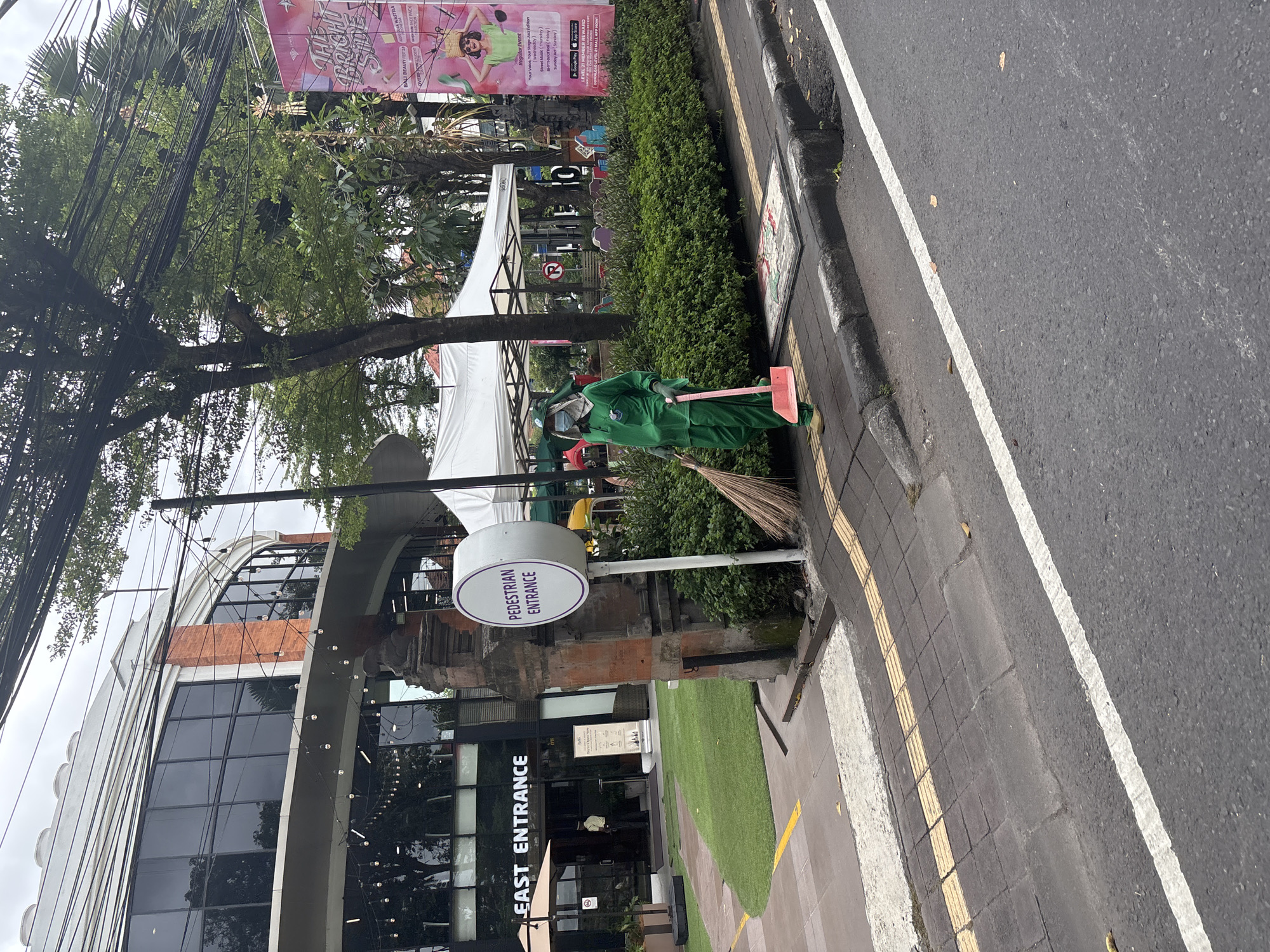A person dressed in a bright green full-body protective suit and face mask is standing on a sidewalk in front of a lush green hedge. They are holding a broom in their right hand and a pink dustpan in their left hand, appearing to be engaged in cleaning. The sidewalk has yellow tactile paving and a gray concrete curb. Behind the person, a white sign with the words "PEDESTRIAN ENTRANCE" in purple lettering is mounted on a rustic stone pillar. Further back, a building with large windows and an "EAST ENTRANCE" sign is visible. A white tent structure is set up in front of the building, and trees with dense foliage are present in the background. A large banner with event details is also visible on the right side of the image. The scene is outdoors, with an overcast sky. The location is identified as Denpasar, Indonesia.