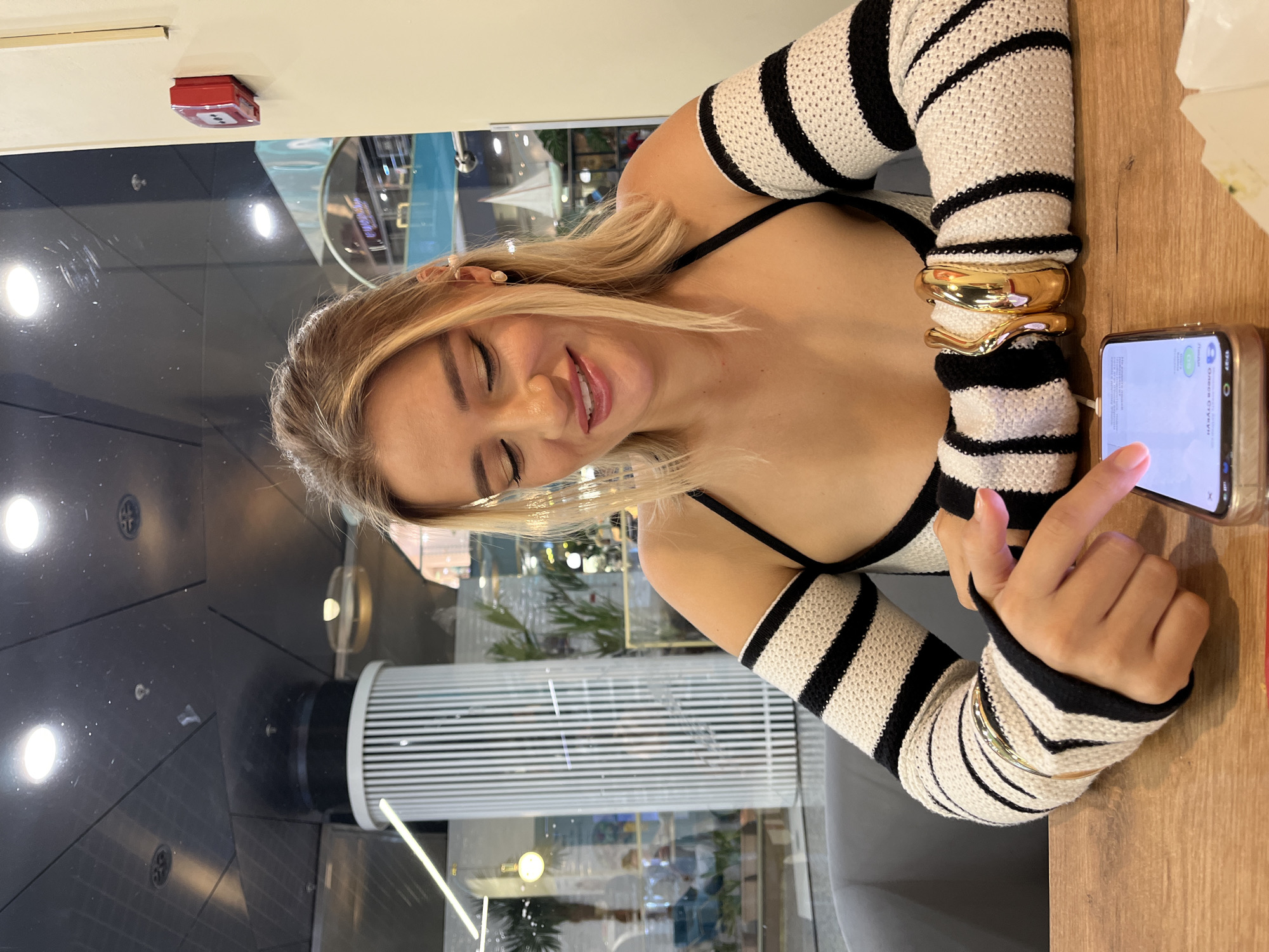 A blonde woman sits at a wooden table, looking down at her phone. She is wearing a black and white striped garment with detached sleeves, and gold bracelets on her left arm. In the background, through a glass window, a cylindrical column with vertical white slats is visible, along with other interior elements of what appears to be a shopping mall or public space. The lighting suggests an indoor setting. The scene is set in Moscow, Russia.