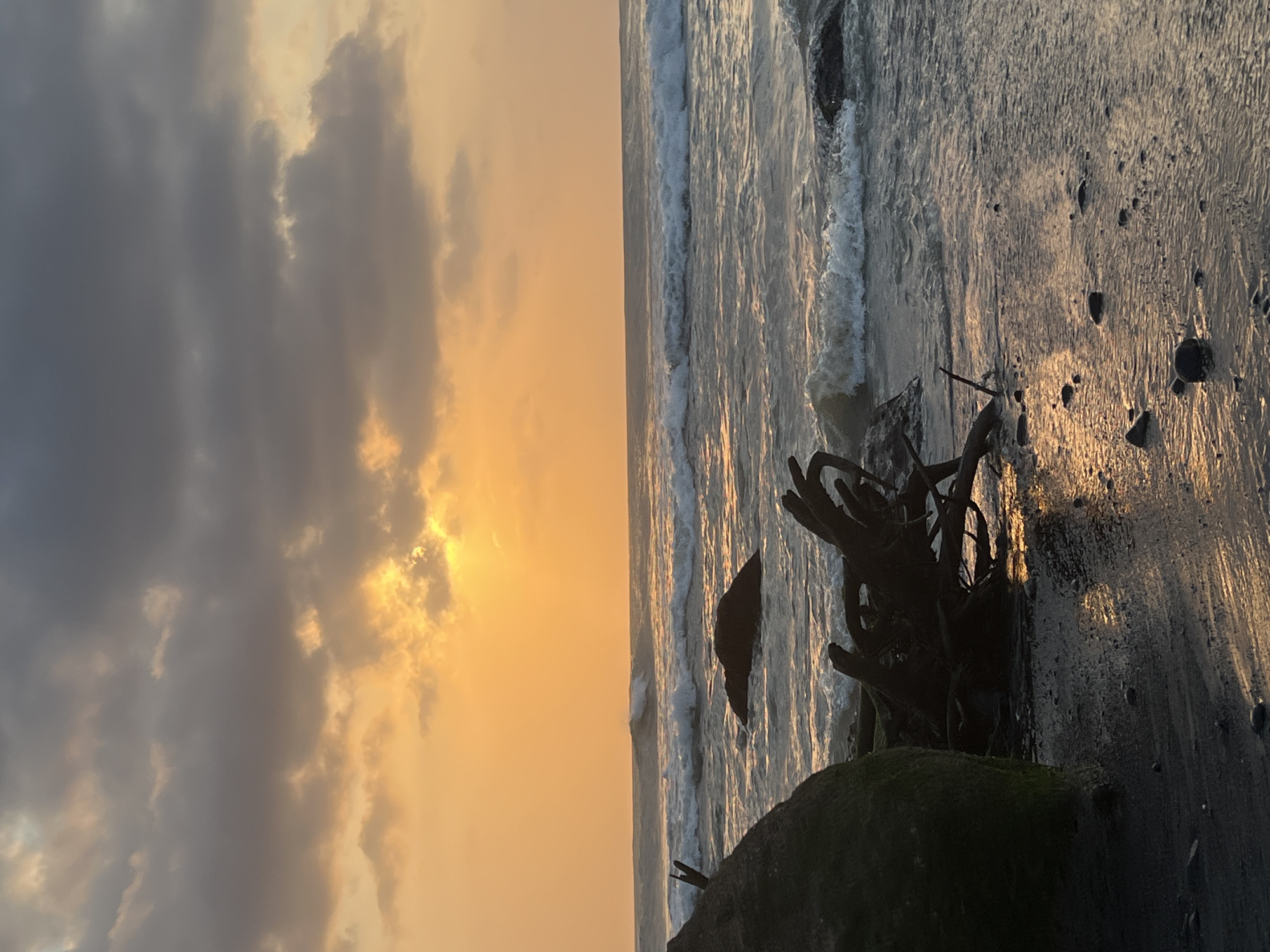 This image depicts a serene beach scene at sunset. The sky is a gradient of warm oranges and yellows, with dark gray clouds partially obscuring the sun. The sun's rays are breaking through, creating a warm glow. Waves gently roll onto a dark sand beach, where driftwood and rocks are scattered. The overall mood is peaceful and tranquil, evoking the beauty of a coastal sunset. There are no people or specific landmarks visible to indicate the location within the city of Kediri, Indonesia.