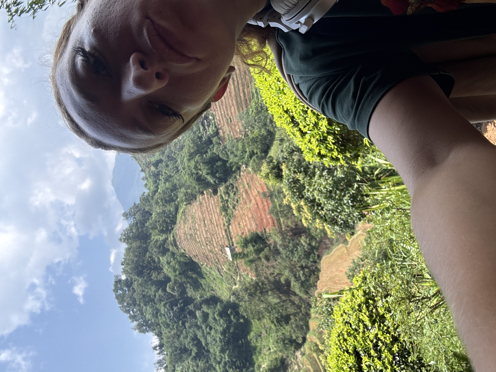 The photograph shows a person taking a selfie. The background is a landscape featuring terraced fields, common in mountainous agricultural areas. Green vegetation covers the slopes, indicating a lush environment. Sky with scattered clouds visible. Location appears to be in the Hoàng Su Phì Commune, Vietnam.