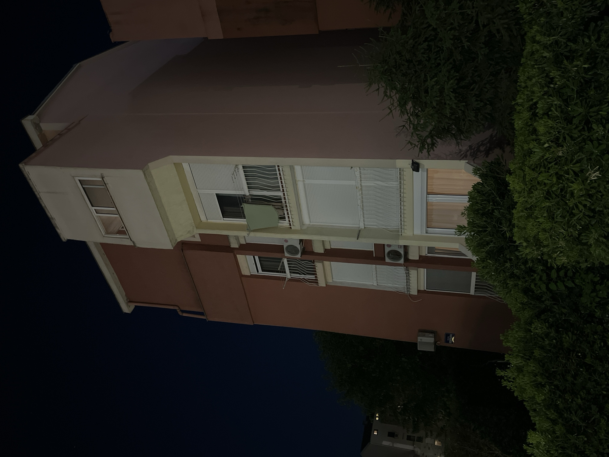 The image shows the side of a multi-story apartment building at night in Budva, Montenegro. The building has a reddish-brown facade and cream-colored trim around the windows and balconies. There are several windows with white blinds, and some balconies with white railings. An air conditioning unit is visible on one of the balconies. A green cloth is hanging on a clothesline on one of the balconies. There are trees and bushes in the foreground and to the sides of the building, and another building with lit windows is visible in the distance to the left. The sky is dark blue, indicating it is nighttime. A street sign is mounted on the reddish-brown facade of the building.