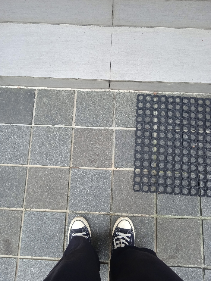 The image shows a person's feet standing on a tiled floor in front of a black rubber mat. The person is wearing black pants and black and white sneakers. The tiles are a light grey color and are arranged in a grid pattern. The rubber mat is in the upper right corner of the image. The location of the image is in Surakarta, Indonesia, at coordinates -7.569 latitude and 110.811 longitude. The photo is taken from a bird's eye perspective, and the person is standing in the middle of the frame. The image is well-lit and the colors are clear.