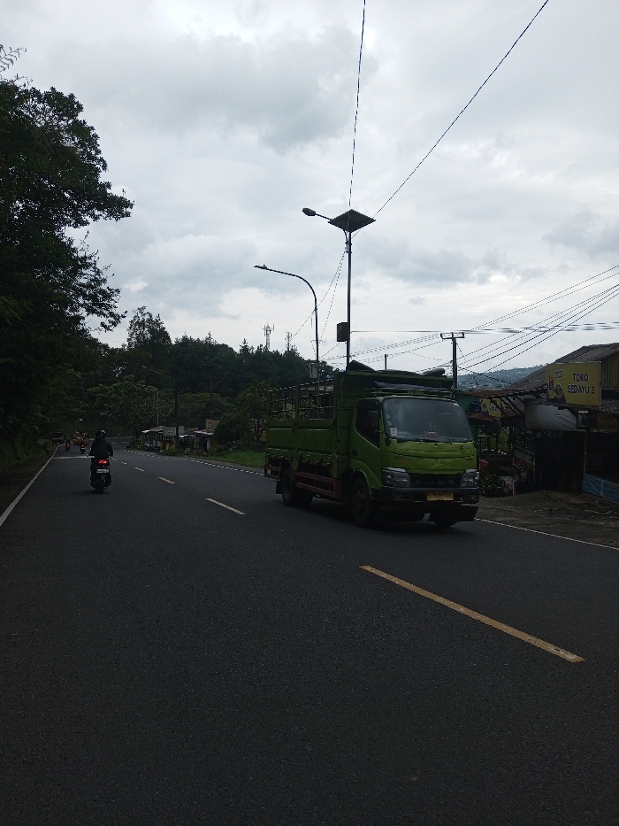 The scene depicts a two-lane asphalt road in Ciherang, Indonesia, under an overcast sky. A light green, medium-duty flatbed truck with a high-sided cargo cage is positioned in the right lane, moving towards the viewer's right. Its front bumper displays a yellow license plate. Further down the road, a solitary motorcyclist, wearing dark attire and a helmet, is operating a motorcycle in the left lane, moving away from the viewer. The road surface features a broken yellow line separating traffic directions and a solid white line marking the right shoulder. To the right of the road, a semi-permanent structure identified by a faded yellow sign reading "TOKO SEDAYU 2" is visible. Multiple utility poles with a dense network of overhead electrical and communication wires are present near the structure. One prominent streetlight post features a solar panel array atop its fixture. The left side of the road is bordered by dense green foliage, transitioning into tree-covered hills in the distance, where telecommunication towers are discernible. The sky is uniformly grey and heavily clouded.