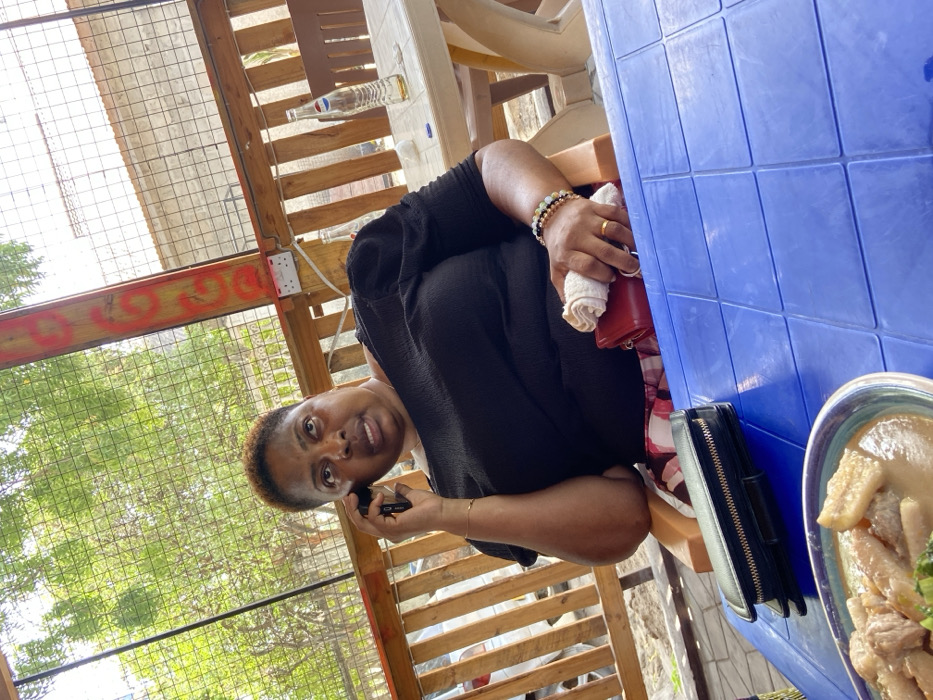 The image shows a woman sitting at a table outdoors in Dar es Salaam, Tanzania, at the coordinates -6.77 latitude and 39.225 longitude. The woman is wearing a black shirt and holding a cell phone to her ear. Her left hand is resting on the table, and she is wearing bracelets on her wrist. She is also wearing a red purse and a black wallet. The table is blue and has a plate of food on it. There is a wooden fence behind the woman, and a green tree can be seen through the fence. The woman appears to be on the phone and looking at the camera. The scene appears to be during the day. The image was likely taken in a local eatery, as the surrounding area suggests a casual, outdoor setting.