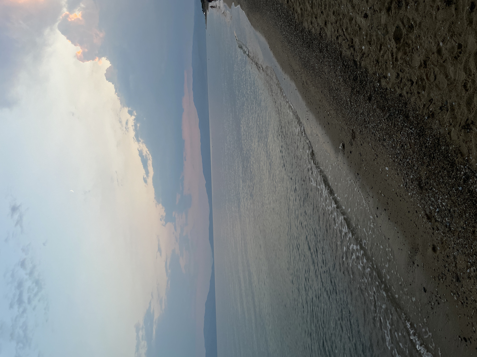 This image depicts a beach scene in Kariani, Greece, during twilight. The sky transitions from a pale blue near the horizon to a soft lavender and pink at the top, with scattered clouds. A sliver of the moon is visible in the upper left quadrant. The sea is calm, with gentle waves lapping the shore. The beach itself is a mix of sand and pebbles, with some faint footprints visible on the sand. In the distance, a silhouetted coastline with mountains can be seen. On the right side of the image, along the waterline, two small figures of people are visible walking.