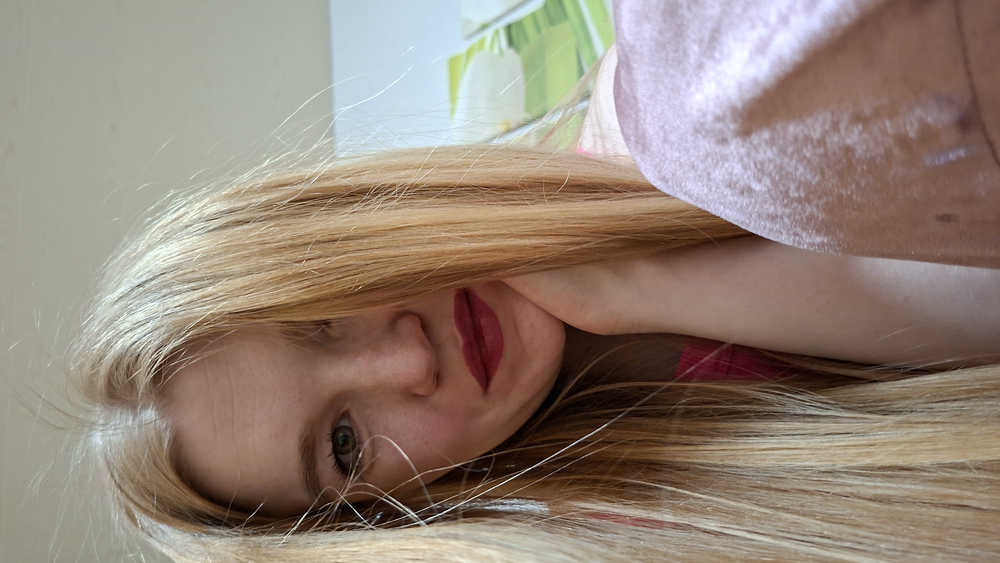 A close-up portrait features a young person, likely female, with long, bright blonde hair cascading around her. She is positioned on her side, resting her head on her hand or arm, and looks directly towards the viewer with one visible green or hazel eye. Her fair skin shows a subtle blush, and her lips are adorned with dark red lipstick.
The blonde hair is a prominent element, appearing fine and reflecting light, framing her face and flowing across a large portion of the frame. In the upper right corner, a soft, pinkish-peach textured fabric, possibly clothing or bedding, is visible. The background is softly blurred but reveals a light-colored wall with faint green and white patterns, suggesting an indoor setting, possibly a home. The lighting is gentle and diffused, casting no harsh shadows. The person's expression appears calm and contemplative. There is no visible text in the image. The scene takes place indoors, in the context of Большой Букор, Russia.