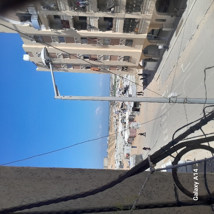 The image, captured from an elevated viewpoint in Khan Yunis, Palestinian Territory, presents a daytime scene under a clear blue sky. In the foreground, dark cables cross the frame, partially obscuring a light-colored building edge on the left. A light gray street light pole stands prominently in the mid-ground, casting a shadow to its left.
Below the street light, a paved road with a pedestrian crosswalk extends horizontally. Individuals are visible on and adjacent to the road; one person walks left-to-right on the crosswalk, another stands near a building to the far right, and a small group moves on the unpaved ground to the right of the road.
To the right of the street light and road, a multi-story residential building with a light-beige facade, balconies, and patterned window surrounds occupies the right side of the frame. Laundry or fabrics hang from multiple windows and balconies, and a white water tank is visible on its roof.
To the left of the street and extending into the background, an expansive area of makeshift shelters, tents, and rudimentary structures is spread across sandy, uneven terrain, indicative of an informal settlement or refugee camp. Several people are observed moving within this area. Beyond the camp, large mounds of earth or sand are present, with the distant Mediterranean Sea visible on the horizon. The scene depicts a populated area blending urban residential structures with an extensive, temporary settlement.