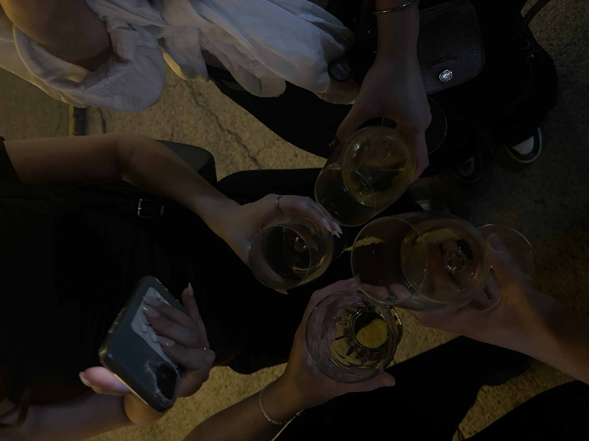 This is a close-up, top-down medium shot of four individuals gathered outdoors at night, likely on a cobblestone street given the texture of the ground. The focus is on their hands holding glasses of amber-colored liquid, suggesting they are toasting or celebrating. One person on the left is holding a smartphone, its screen illuminated. Another person in the center-right is wearing a white shirt. The individuals appear to be in Novi Sad, Serbia, though the specific location within the city is not discernible from the image alone. The overall mood is convivial and social.