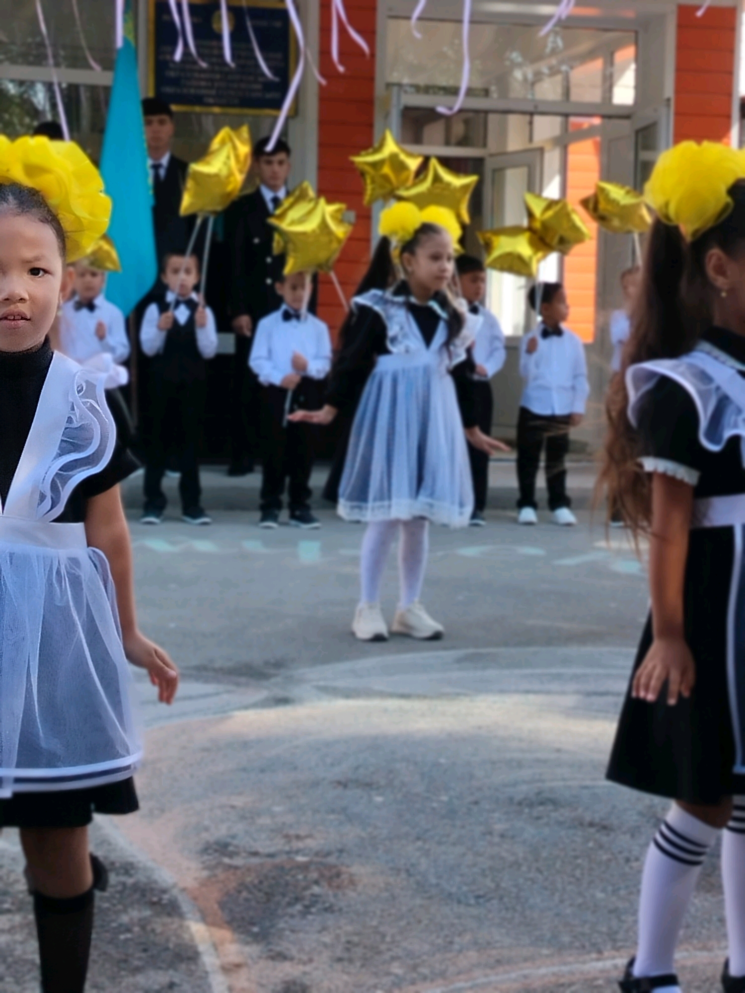 In the city of Kölkent, Kazakhstan, a group of schoolchildren are gathered outdoors, likely for a school event. The foreground features two young girls in traditional school uniforms, characterized by black dresses with white pinafores and yellow hair accessories. The girl on the left is partially visible, facing the camera with her head tilted slightly. The girl on the right is also visible, with her face turned away from the camera, and she is holding a bouquet of golden star-shaped balloons. Behind them, several other children and adults are present, some holding similar balloons. The background shows a building with orange and white accents, and a blue flag, likely the flag of Kazakhstan, is prominently displayed. The ground appears to be asphalt, with some chalk markings visible. The overall atmosphere suggests a festive occasion, possibly the beginning or end of the school year.