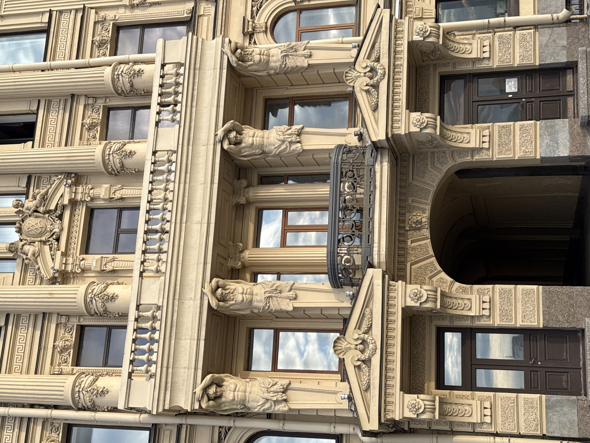 The image shows the facade of a historic building in Saint Petersburg, Russia, likely a palace or a grand apartment building. The architectural style is ornate and classical, with a prominent beige stone facade. Key features include large Corinthian columns, intricate carvings, decorative reliefs, and a balcony with a stone balustrade. There are two large statues of Atlantes holding up the upper structure, located on either side of a central balcony. The windows are tall and rectangular, with some reflecting the sky and clouds. Below the balcony, there is an arched entryway leading into the building. The entrance is flanked by decorative pilasters and topped with a pediment. The overall impression is one of grandeur and historical significance, typical of Saint Petersburg's imperial architecture.