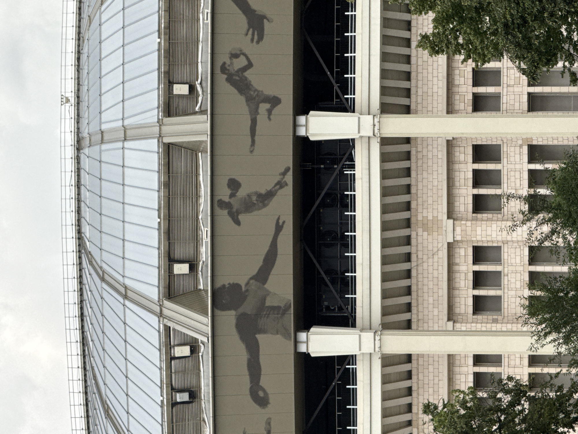 The image depicts the exterior of the Luzhniki Stadium in Moscow, Russia. The stadium features a distinctive domed roof constructed of glass panels. Below the roof, a beige-colored band runs along the exterior, adorned with large, dark silhouette images of athletes engaged in various sports, including what appears to be discus throwing and basketball. The stadium's facade is characterized by large, pale stone blocks and rows of windows. Trees and foliage are visible in the foreground, suggesting an outdoor setting. The sky is overcast.
