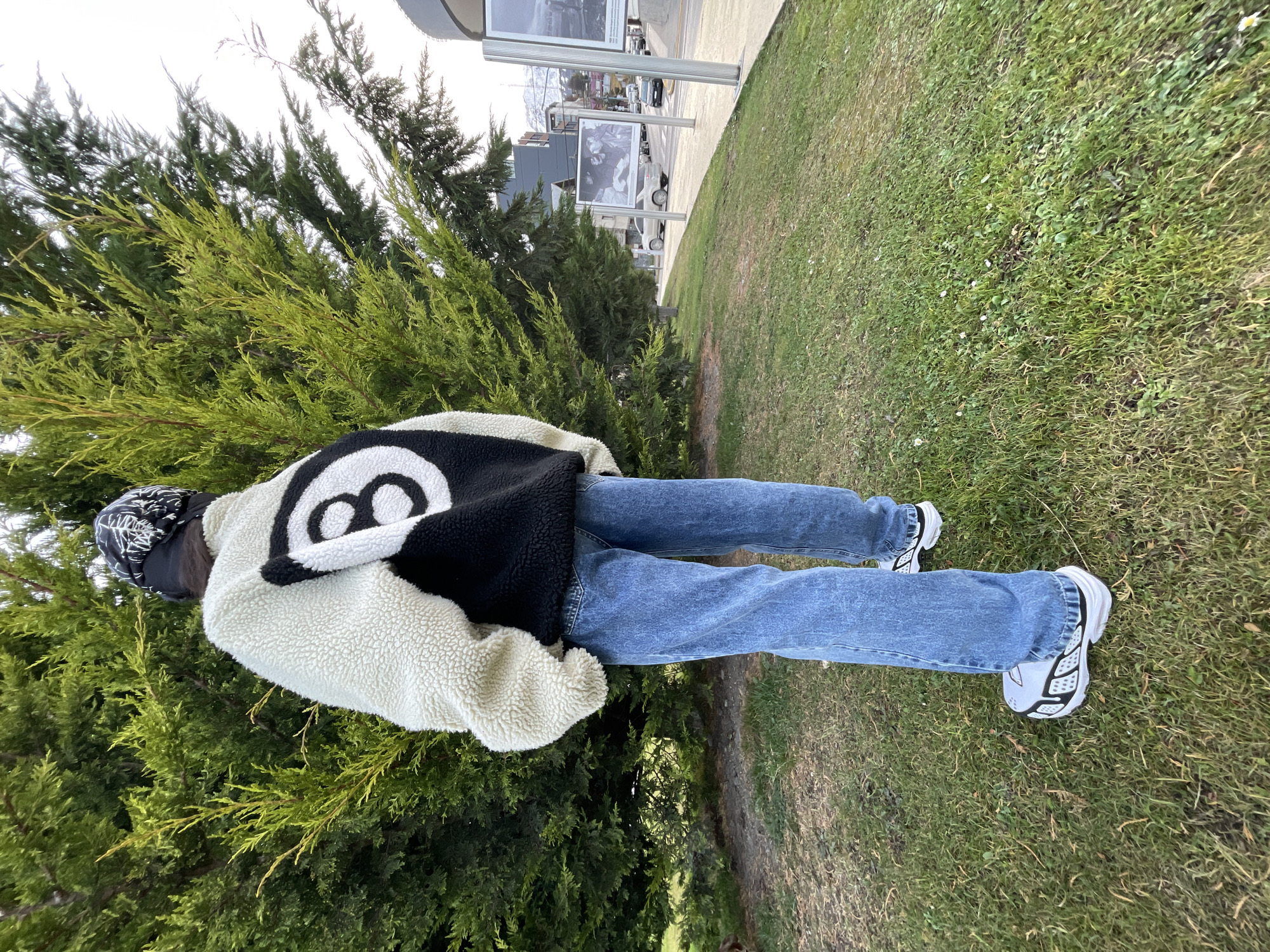 The image shows a person standing on a grassy area with a tree to their left. The person is wearing a white and black jacket with a large "8" ball design on the back, blue jeans, and white sneakers with black detailing. They are also wearing a black and white bandana. The location appears to be Ushuaia, Argentina, judging by the presence of signage visible in the background. The person is facing away from the camera and towards the signage and the street in the distance. The scene appears to be outdoors on a partly sunny day.