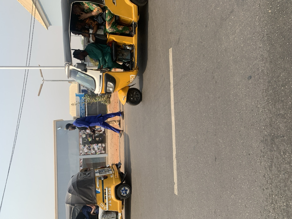 This image captures a street scene in Adankolo, Nigeria, under a clear, bright sky. The foreground features an asphalt road with a faded white painted line. Two yellow motorized tricycles, known as Keke Napeps, occupy the road.
On the right, a Keke Napep is in motion, moving from right to left. Its male driver, wearing a dark green traditional outfit, is visible at the controls. The passenger compartment contains at least three individuals; a female passenger in a green, orange, and black patterned traditional dress is prominent, with her right hand raised near her face.
On the left, another yellow Keke Napep is partially visible from the rear, either stationary or moving slowly away from the viewer. Its rear plate area displays "A-321". At least two passengers are discernible within its canopy.
Between the two vehicles, on a dirt shoulder adjacent to the road, a male pedestrian walks from right to left. He is dressed in a royal blue traditional Nigerian garment. The lower legs of another person are partially visible behind him.
In the background, a light blue building with a black roof and multiple windowed storefronts is visible on the left. Further behind and to the right, a second, lighter blue building with a cream roof is present. A tall, light-colored street lamp pole with two horizontal fixtures stands near the center-right, extending upwards. Multiple dark overhead utility lines span across the upper portion of the frame. The distant sky appears light blue and slightly hazy.