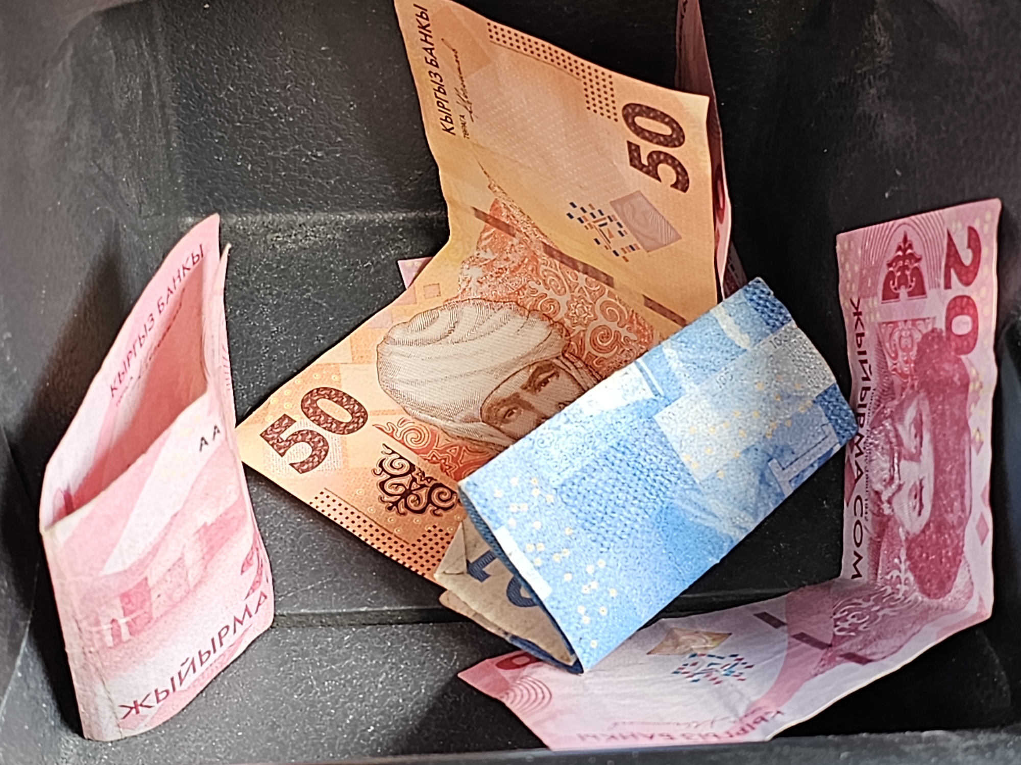 The image shows a close-up of several Kyrgyzstani banknotes, varying in color and denomination, arranged within a dark, textured compartment, possibly a car's center console or a small storage box. The denominations visible are 50, 20, and a partial view of what appears to be a 100 or 200 som note based on the color and portrait. The notes are folded and overlapping, with some partially obscured. The environment is indoors and dimly lit, focusing attention on the currency. There are no people or specific city landmarks visible. The image is a still life of money.