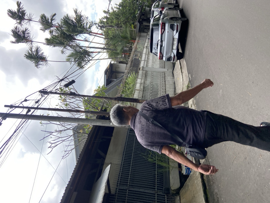 The image shows a man walking on a paved road in Bandung, Indonesia, at the coordinates -6.939 latitude, 107.572 longitude. He is wearing a black t-shirt with "Chanel Paris" printed on it, black pants, and a black shoulder bag. He is walking away from the camera and towards the right side of the frame. There is a silver car parked on the right side of the road, facing the camera. The car is parked behind the man and is partially obscured by him. The background shows several palm trees and a residential building with a fence. The scene is overcast, and there are power lines running overhead.