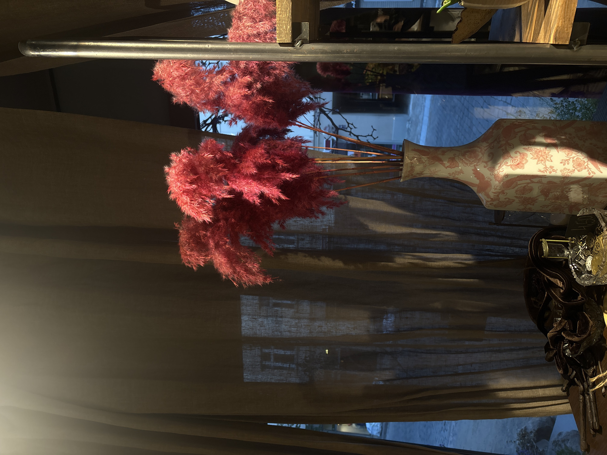 The image depicts a dimly lit interior scene with a window. Through the window, a street scene is visible with buildings and some snow on the ground, suggesting a winter setting. The primary focus in the foreground is a decorative arrangement. A tall, square vase with a pink floral and bird pattern holds a bouquet of dried, fluffy, bright red reeds or pampas grass. To the left of the vase, a dark brown velvet cloth is draped over some dark, metallic objects that appear to be decorative spikes or extensions. A crystal decanter filled with amber liquid and a glass are also present on the surface. To the right of the vase, a vertical metal pipe and wooden shelves are visible, part of a shelving unit. The lighting suggests either early morning or late afternoon sunlight filtering through a sheer, dark brown curtain. The scene appears to be an indoor space, possibly a shop or a home, with a view of a street in Riga, Latvia.