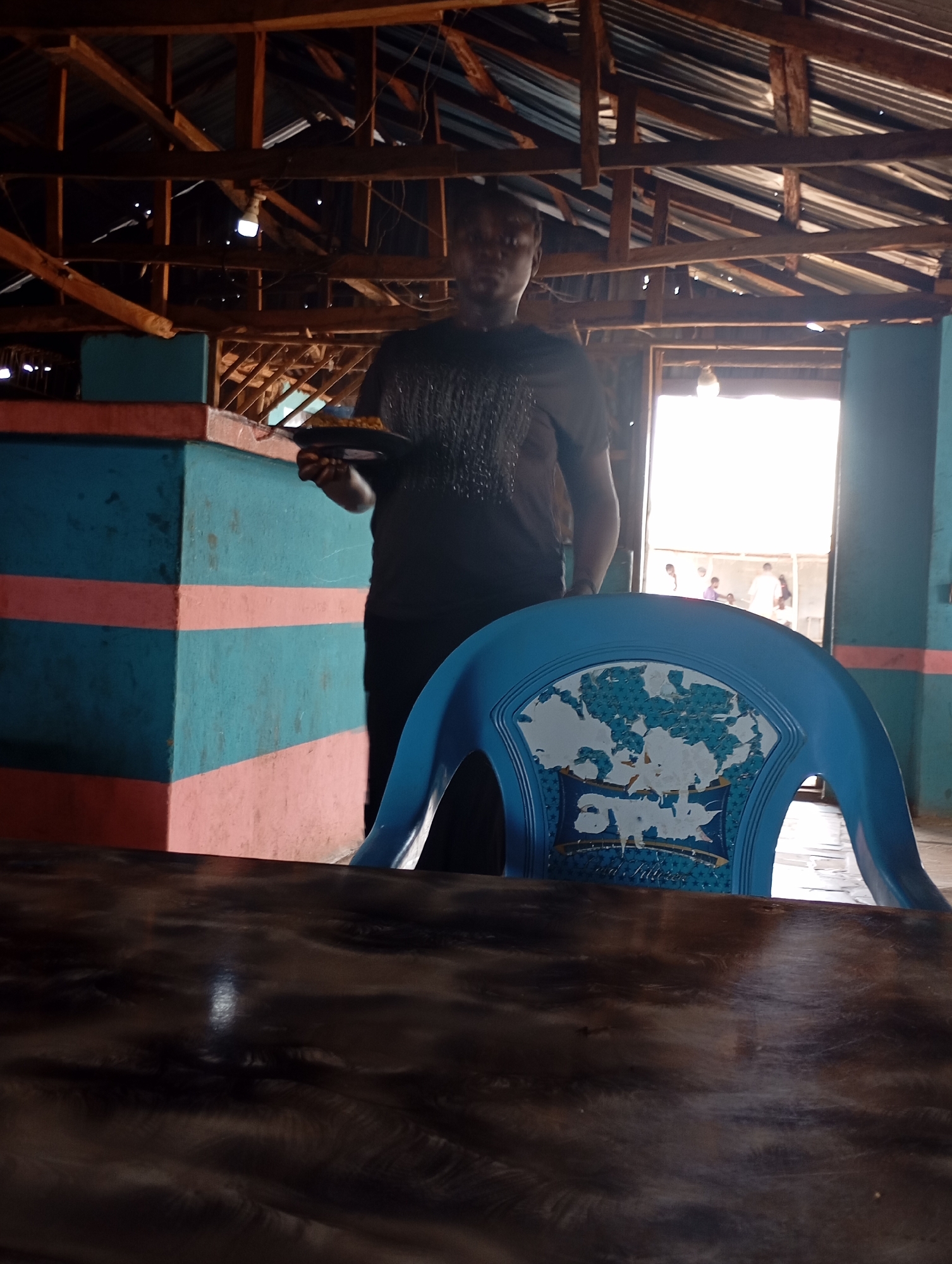 The image depicts an indoor scene, possibly a cafe or restaurant, with a partially visible structure of wooden beams and corrugated metal roofing overhead. The walls are painted with distinct bands of teal and pink. In the foreground, a dark, polished wooden surface, likely a table, occupies the bottom portion of the frame. Behind this, a blue plastic chair with a distressed design on its back is visible.
A person wearing a black t-shirt stands in the midground, holding a dark plate with food. The person's face is not clearly visible due to lighting and angle. Beyond the person, an open doorway leads to an exterior area where several individuals can be seen, silhouetted against bright light.
The overall impression is of a casual, possibly rustic establishment. The location in Shendam, Nigeria, suggests a West African context. The presence of food and people in the doorway indicates ongoing activity. The worn appearance of the chair and the exposed roofing suggest an aged or utilitarian setting.