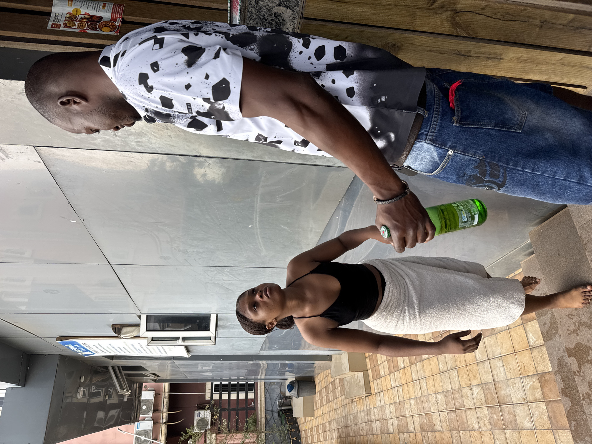 In Nkpor, Nigeria, on a tiled walkway leading up to a building with a grey facade and wooden accents, a woman and a man are interacting. The woman, wearing a black halter top and a white towel wrapped around her lower body, stands on a step looking up at the man. She is barefoot. The man, dressed in a black and white patterned shirt and blue jeans with a red cloth tucked into a back pocket, holds a green bottle in his right hand. The background includes an outdoor air conditioning unit, a trash bin, and a sign on the building.