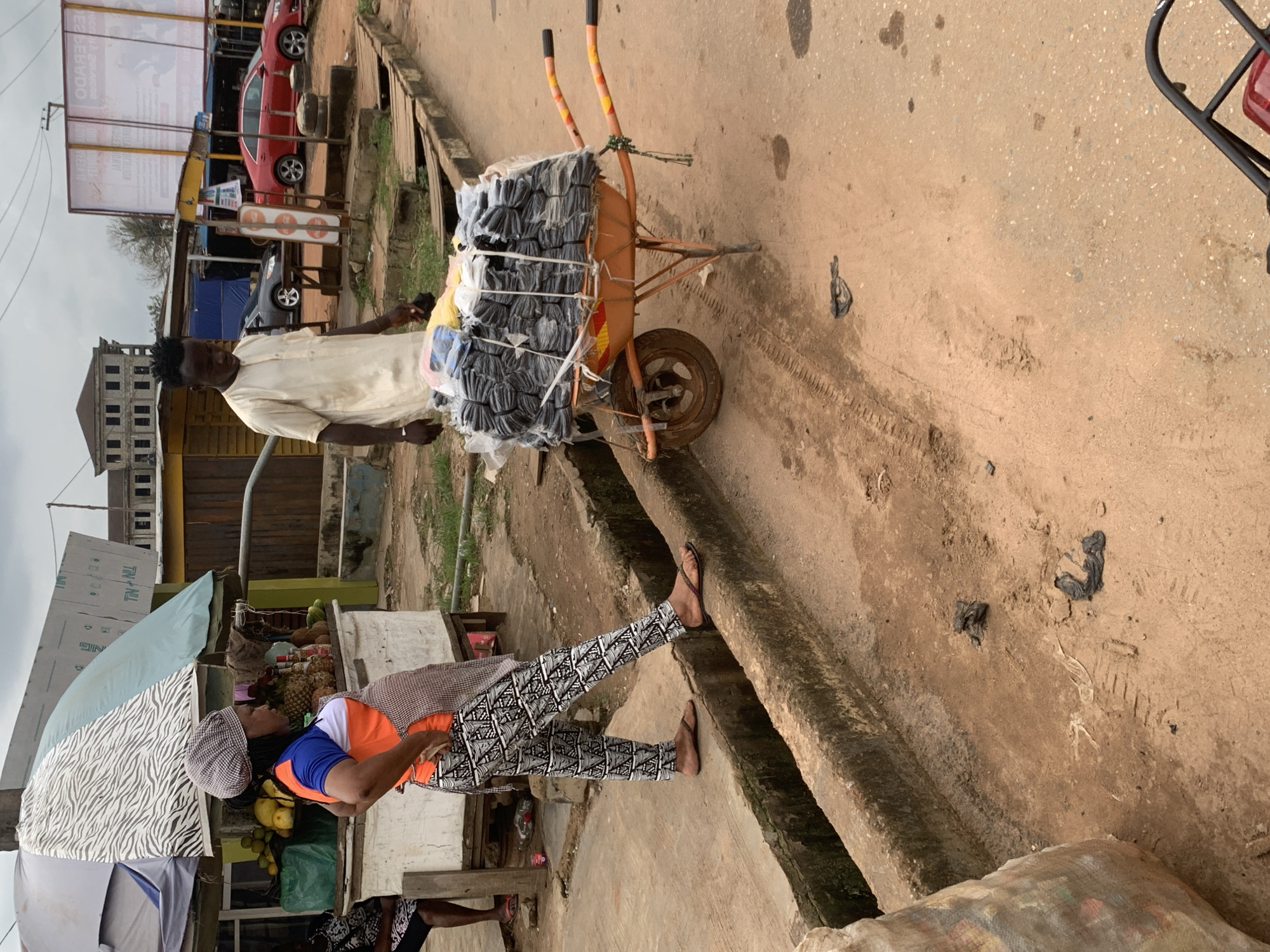 In Shagamu, Nigeria, on a dusty road with a concrete drainage channel, a woman wearing a patterned headwrap, an orange and blue shirt, an apron, and black and white patterned pants stands with her hands on her hips. To her right, a man in a cream-colored robe pushes a wheelbarrow loaded with neatly stacked black items, possibly textiles or packaged goods. The wheelbarrow is orange with yellow and black handles and one visible wheel. In the background, a roadside market stall displays fruits like pineapples and mangoes under a striped umbrella. Several cars are parked along the road, including a red sedan and a grey sedan. A large billboard with text in an unknown language is visible in the distance. The ground is a mix of dirt and some debris, with tire tracks visible in the dust.