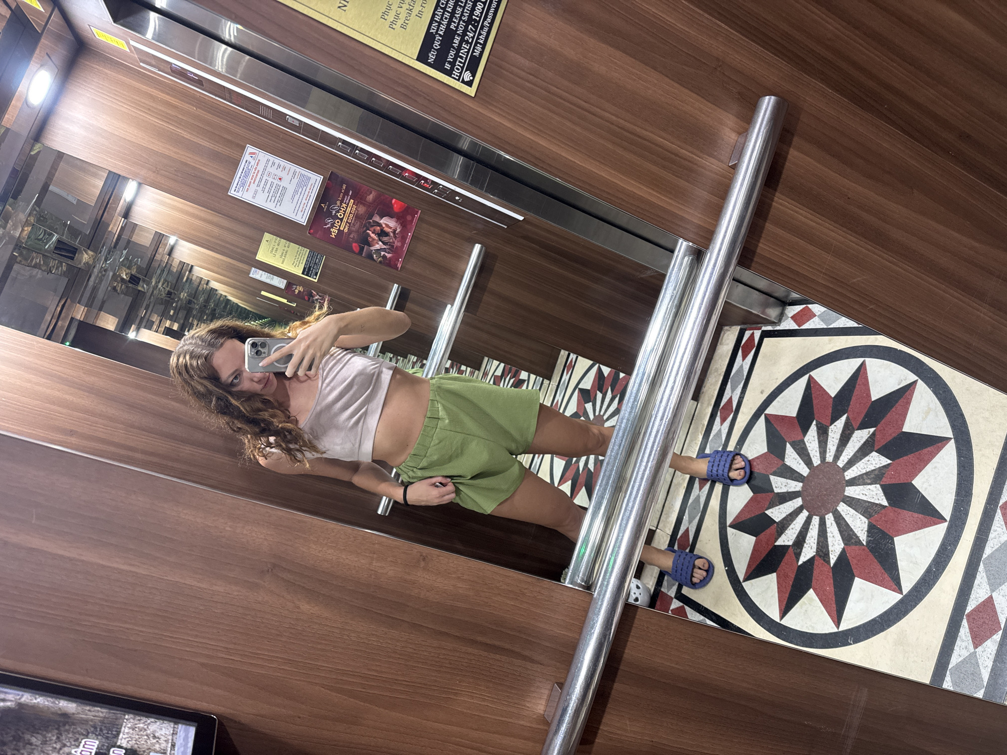 An individual is taking a mirror selfie inside an elevator. The individual is a woman with curly brown hair, wearing a light pink crop top and green shorts. She is holding a smartphone in her right hand, capturing her reflection. The elevator interior features wood-paneled walls, a polished metal handrail, and a decorative floor tile with a starburst pattern. Posters are visible on the elevator walls, including one with text in Vietnamese that reads "NỮO OHN 000,002" and another with "HOTLINE 24/7: 1900". The presence of Vietnamese text and currency notation suggests the location is in Vietnam. Considering the context provided by the text on the posters and the typical branding found in hotels in Hanoi, it is highly probable that this elevator is located in a hotel in Hanoi, Vietnam.