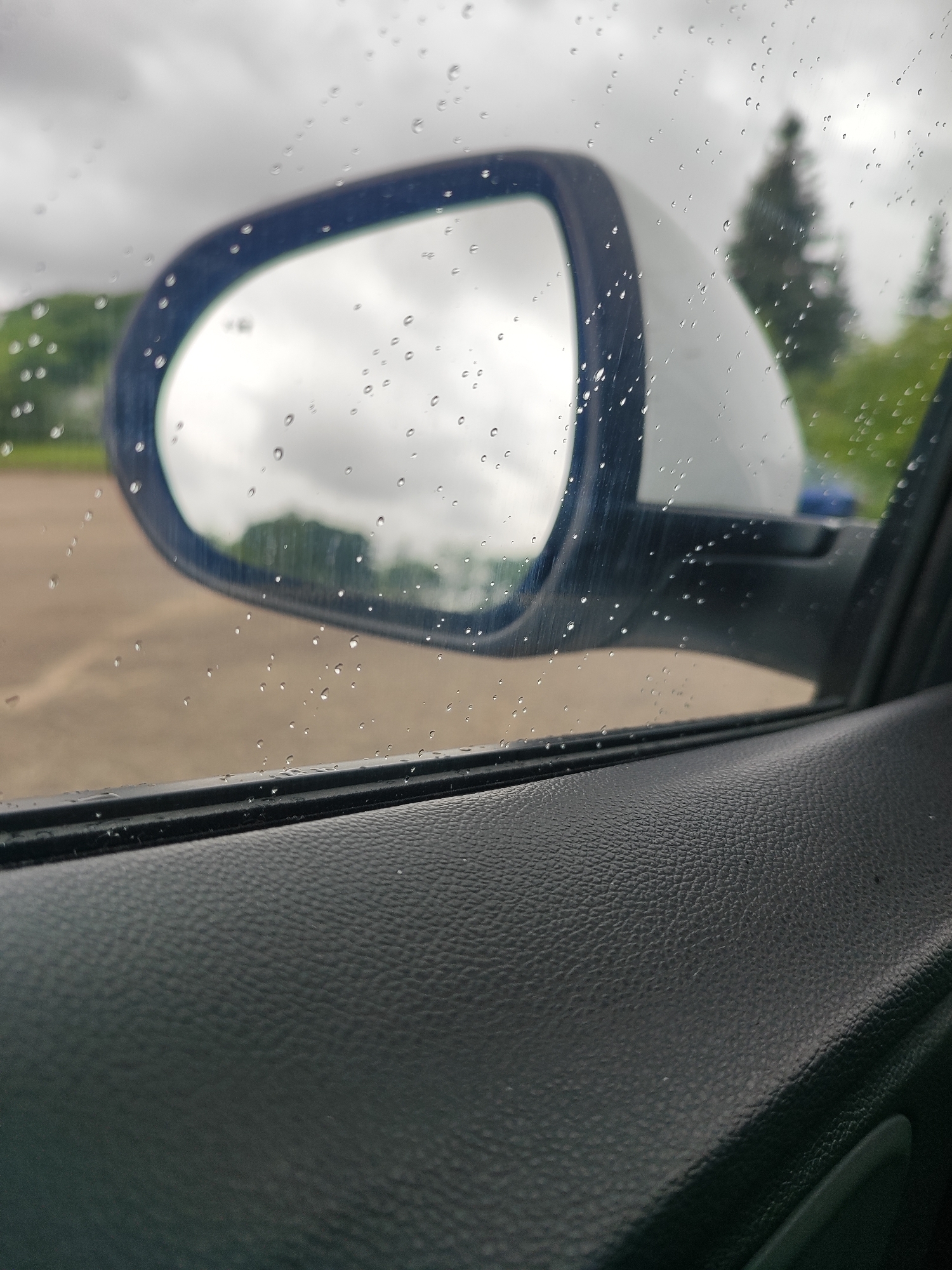 The image captures a close-up view of a car's side mirror and door panel on a rainy day, with water droplets visible on the mirror and window. The car appears to be parked outdoors, evidenced by the blurred background of a paved area and vegetation, including trees and green foliage. The sky is overcast, suggesting a cloudy or rainy weather condition. The interior of the car is partially visible, showing a textured dark material on the door panel. The side mirror has a blue trim and reflects the cloudy sky and distant trees. The scene is likely in Hrodna, Belarus, given the contextual information. There are no people or discernable human activity in the image.